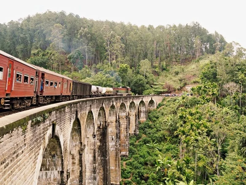 A train with red carriages travels along the Nine Arch Bridge in Ella Sri Lanka