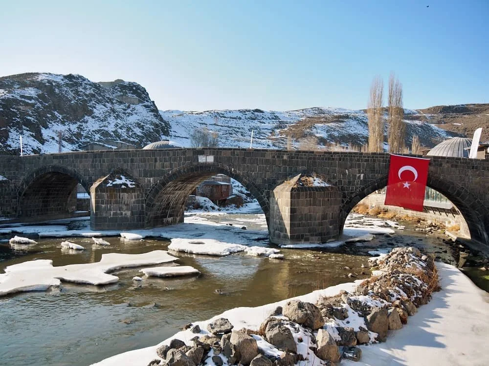 Taş Köprü  over a partially frozen Kars River, with a Turkish flag hanging down from one arch and mountains in the background