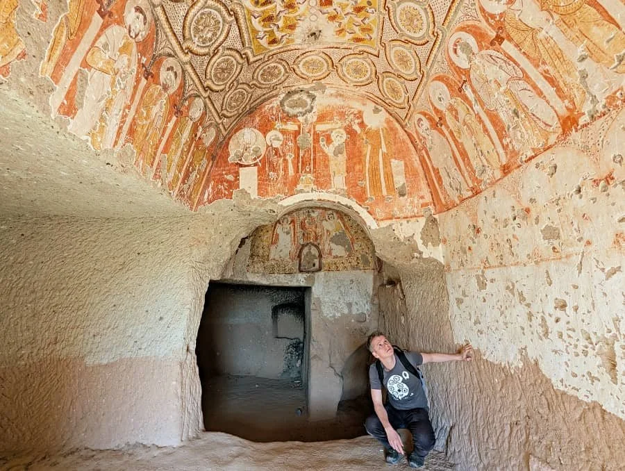 A man in a t-shirt looks up at brightly coloured frescoes in a cave church in Cappadocia, Turkey