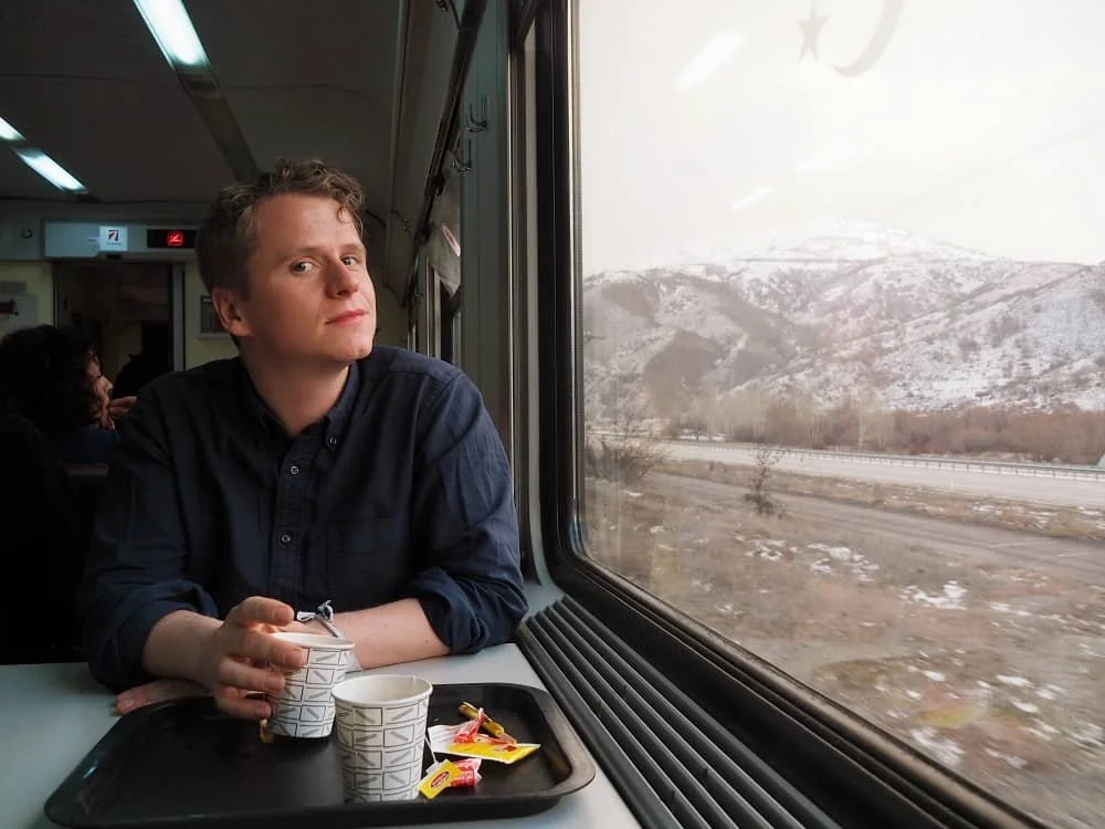 Vincent with a paper cup in his hand in the dining car of the Eastern Express (Dogu Ekspresi) train. He is next to a window outside of which snow covered mountains can be seen.