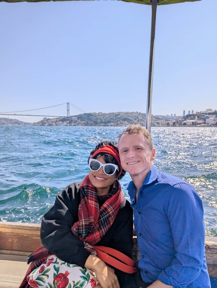 a woman and a man smile for a photo on the small wooden boat with the bosphorus and the bosphorus bridge in the background