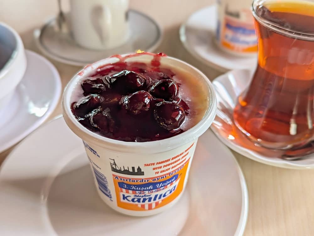 Close up of a pot of kanlica yogurt topped with cherry jam, with a glass of tea in the background