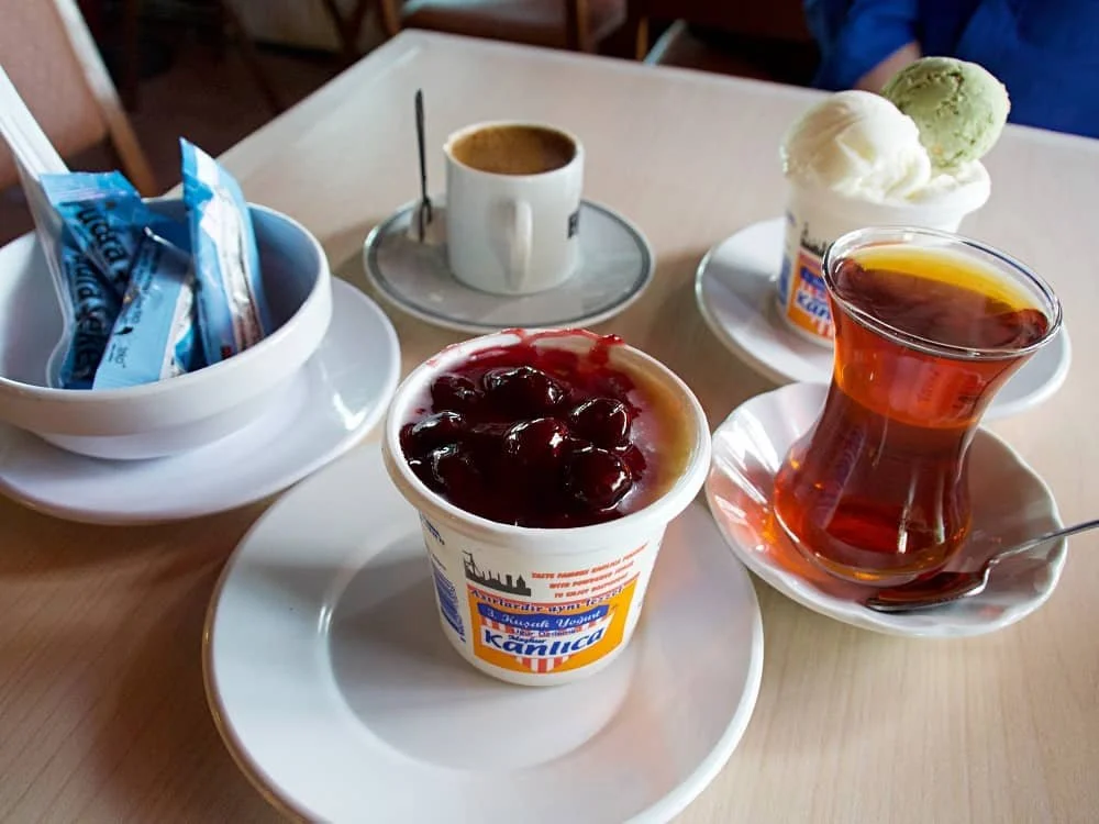 A table with a pot of Kanlica yogurt topped with cherry jam, another pot in the background topped with ice cream, a glass of Turkish tea and a cup of Turkish coffee and a bowl filled with sachets of powdered sugar.