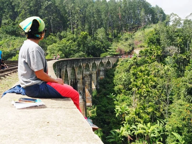 A woman (Sasha) wearing a green bandana, wearing a grey t-shirt and orange trousers sits on the edge of the Nine Arch Bridge in Ella Sri Lanla
