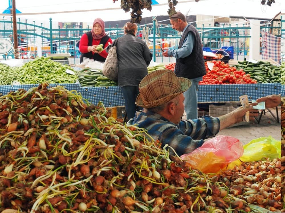 14 day travel itinerary turkey A man in a checked shirt and trilby hat and sitting in front of a pile of shallots exchanges money with a customer at Fethiye Tuesday Market in TĂŒrkiye
