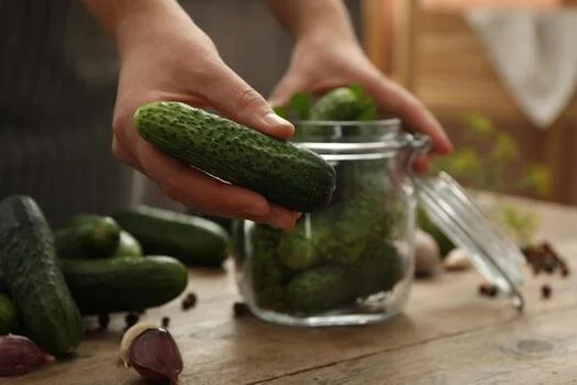 woman-putting-cucumber-into-glass-jar-at-wooden-kitchen-table-closeup-pickling-vegetables-photo.jpg