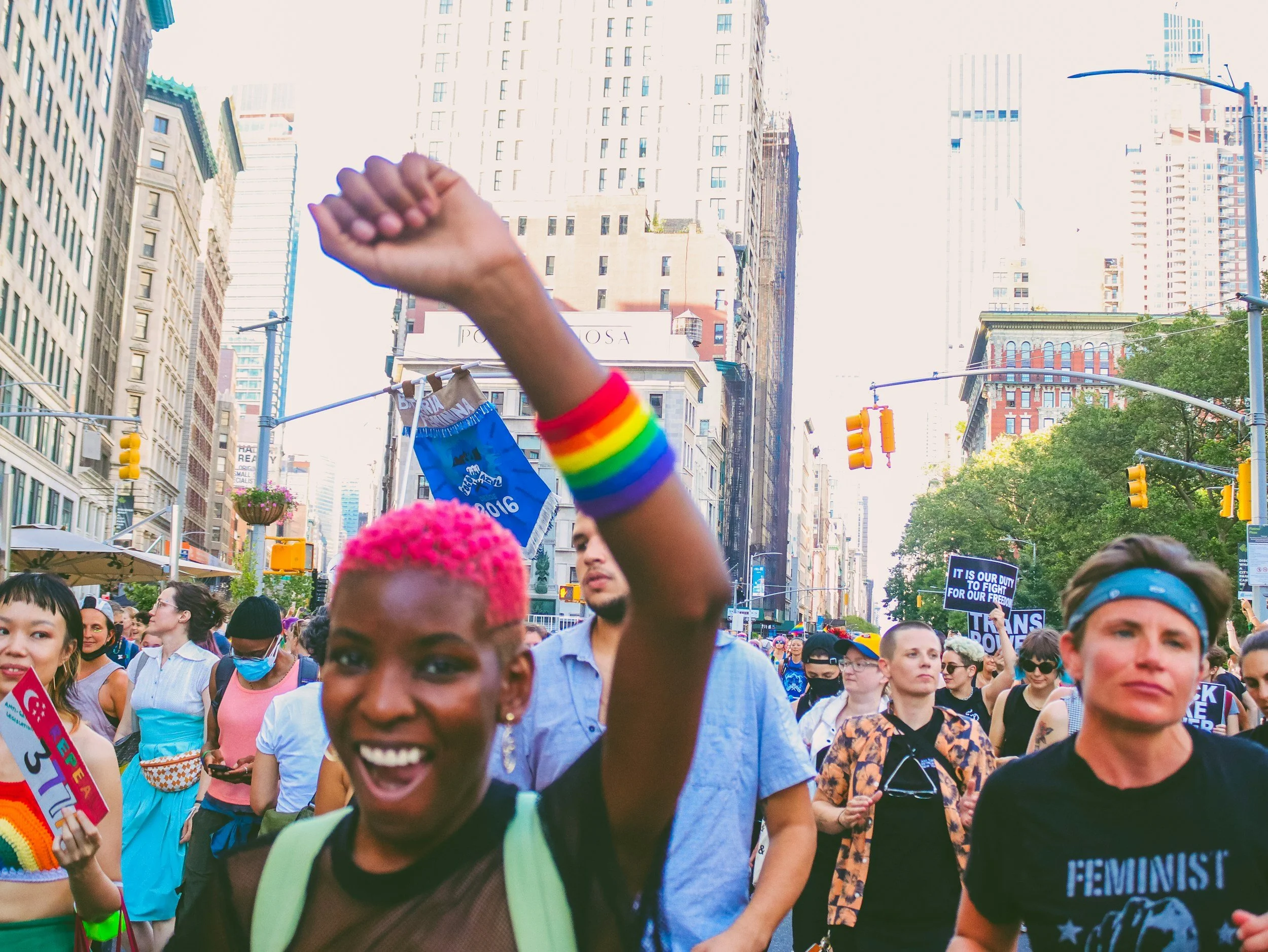People participating in a pride march on a city street, with rainbow accessories and signs, surrounded by tall buildings and traffic lights.