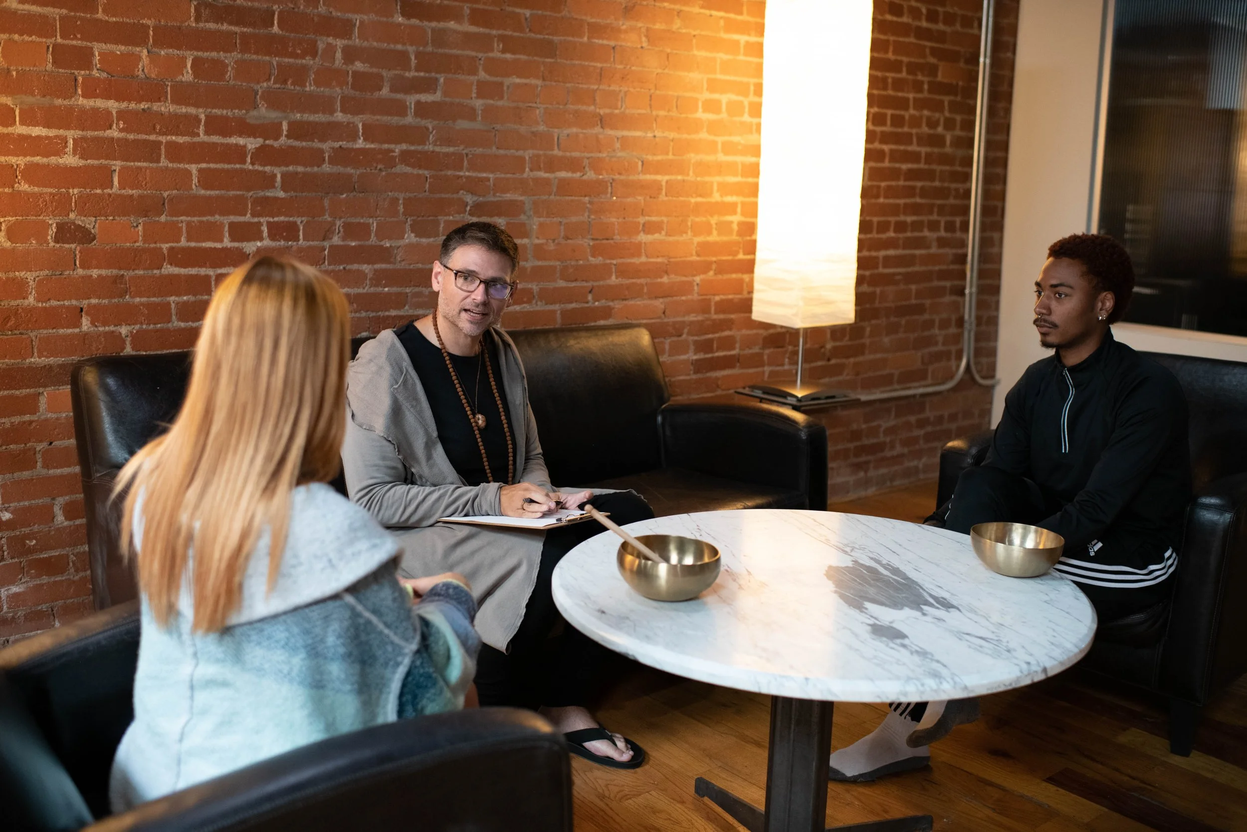 Three people sitting around a round marble table having a discussion in a room with brick walls and a modern lamp.
