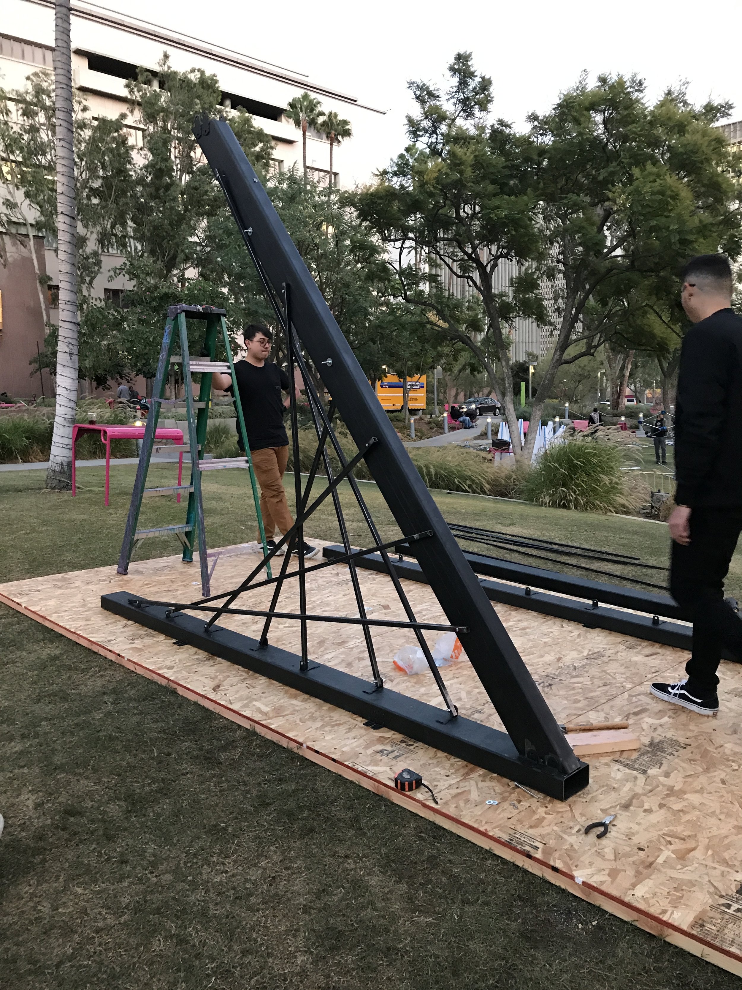 Two people setting up a large black metal structure on a wooden platform outdoors in a park, with trees and a building in the background.
