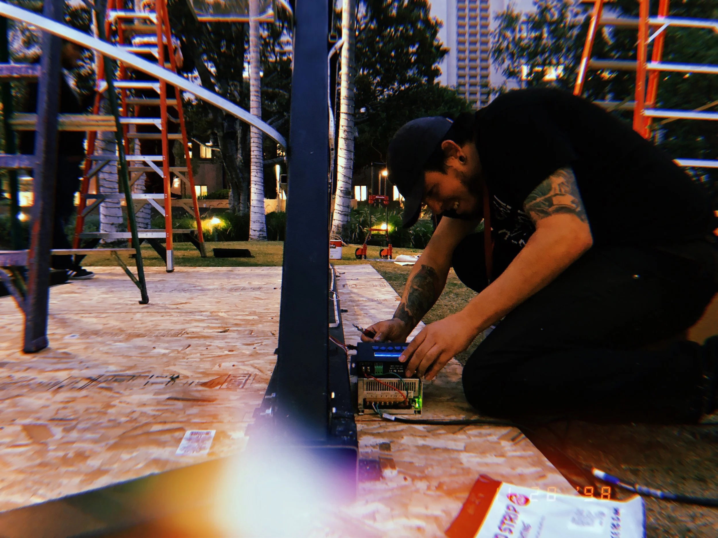 A man with tattoos on his arms is kneeling on the ground, working with electronic equipment near a black metal structure outdoors in a park-like area. There are trees, buildings, and playground equipment in the background.