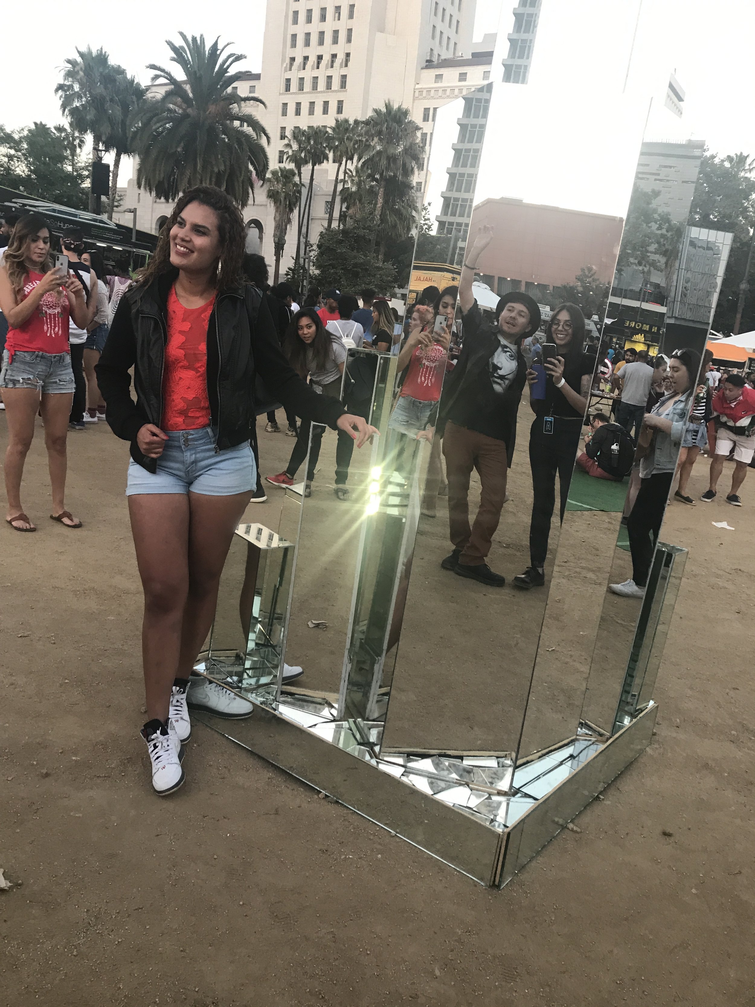 Group of people taking selfies around a large, reflective mirror at an outdoor event in an urban area with tall buildings and palm trees