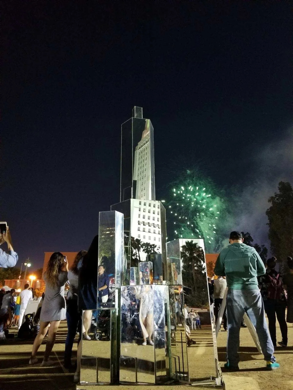 Night scene with a group of people gathered around a tall, reflective, abstract sculpture resembling a skyscraper. Fireworks are visible in the dark sky behind the sculpture, illuminating the scene.