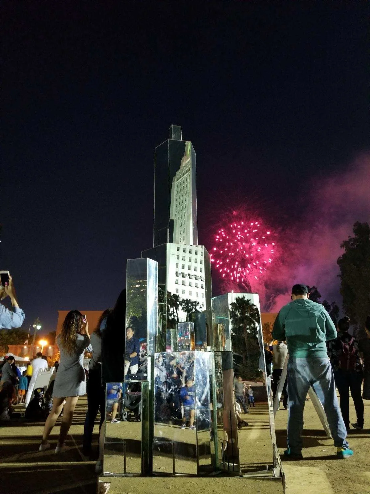 People gathered at night with fireworks in the sky behind a tall building and reflective sculptures in the foreground.