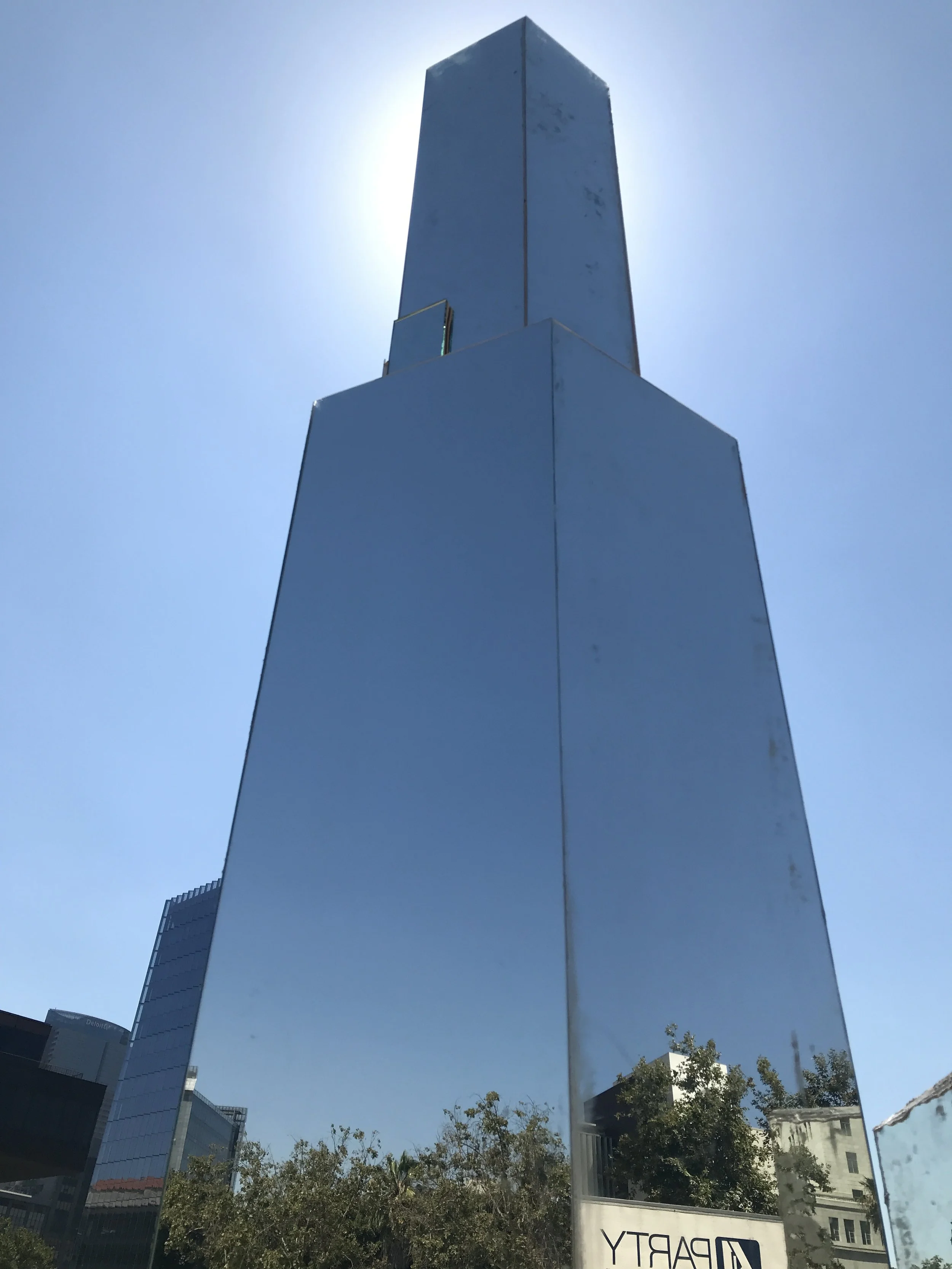 A tall, reflective, blue glass building with a bright sun behind it, reflecting the sky and nearby trees, with a cityscape and trees at the base.