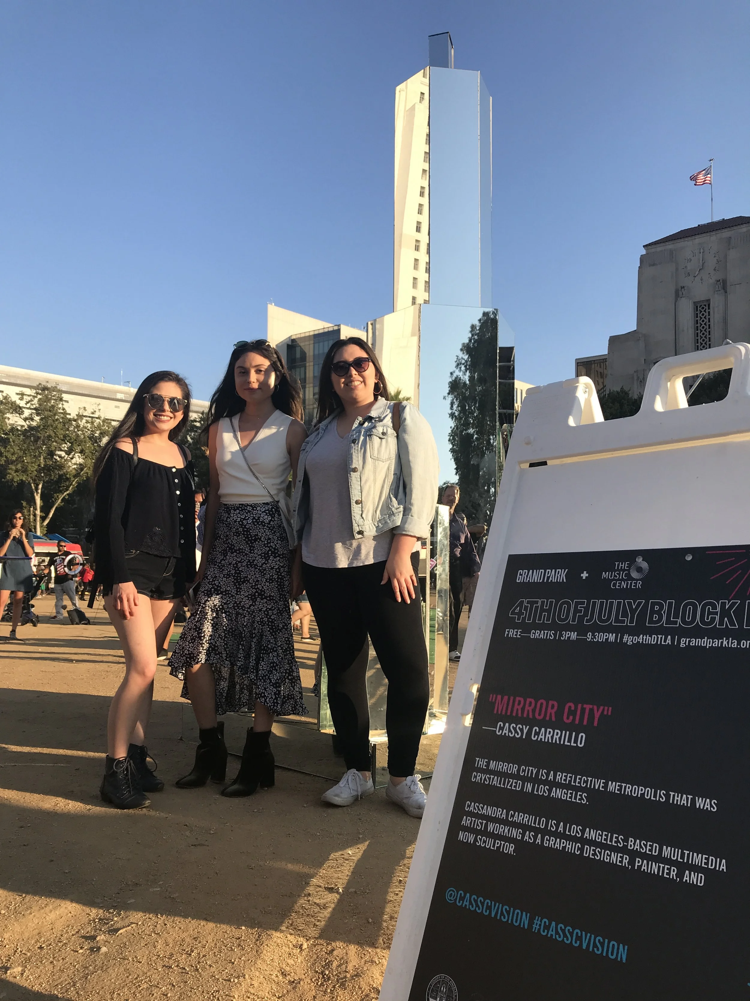 Three women standing outdoors near a reflective skyscraper, smiling at the camera, with an event signboard in the foreground celebrating the 4th of July in Los Angeles.
