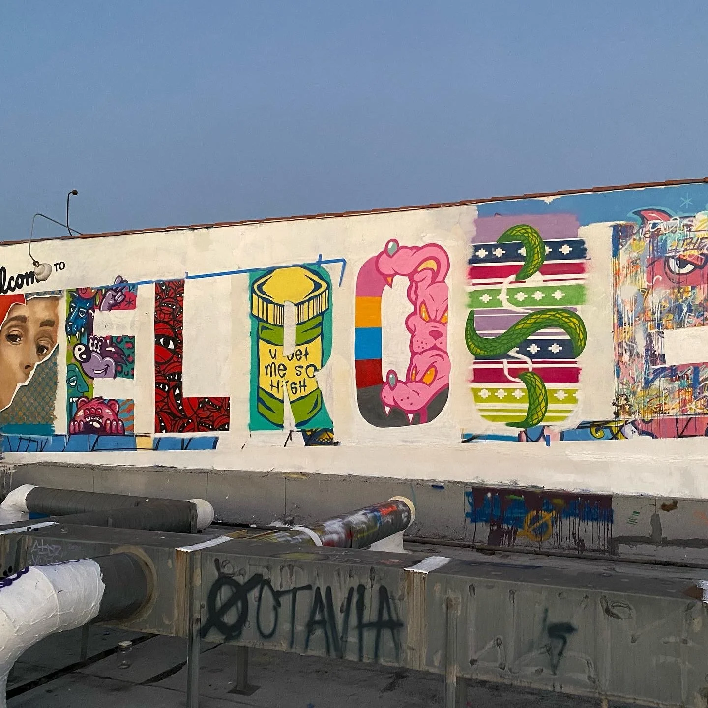Colorful mural on a white wall featuring animated characters and patterns, including a woman’s face, abstract eyes, a can with the words 'Wet me so high,' a pink vinyl record with a snake, and other stylized imagery, with spray paint cans in the fore
