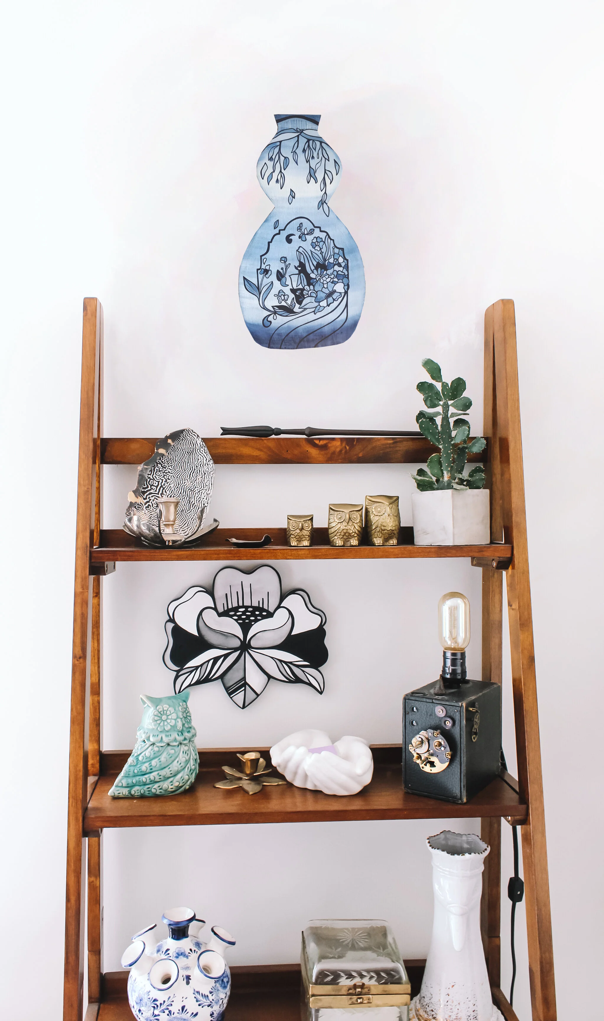 Picture of a shelf with many objects and art on it including the Painting on a vase space, painting is of many colours of blue and an archway with flowers spilling out of it. 