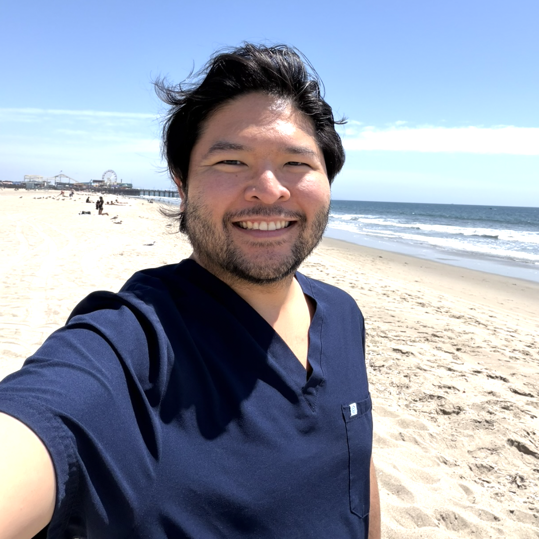 A Japanese man is taking a selfie on a beach. He is wearing blue scrubs and has dark hair and a light beard. 