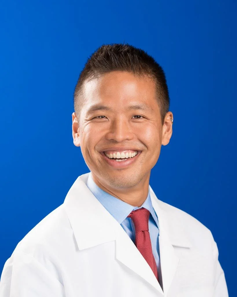 A headshot of a smiling James Huang wearing a white lab coat with a visible blue collared shirt and red tie.