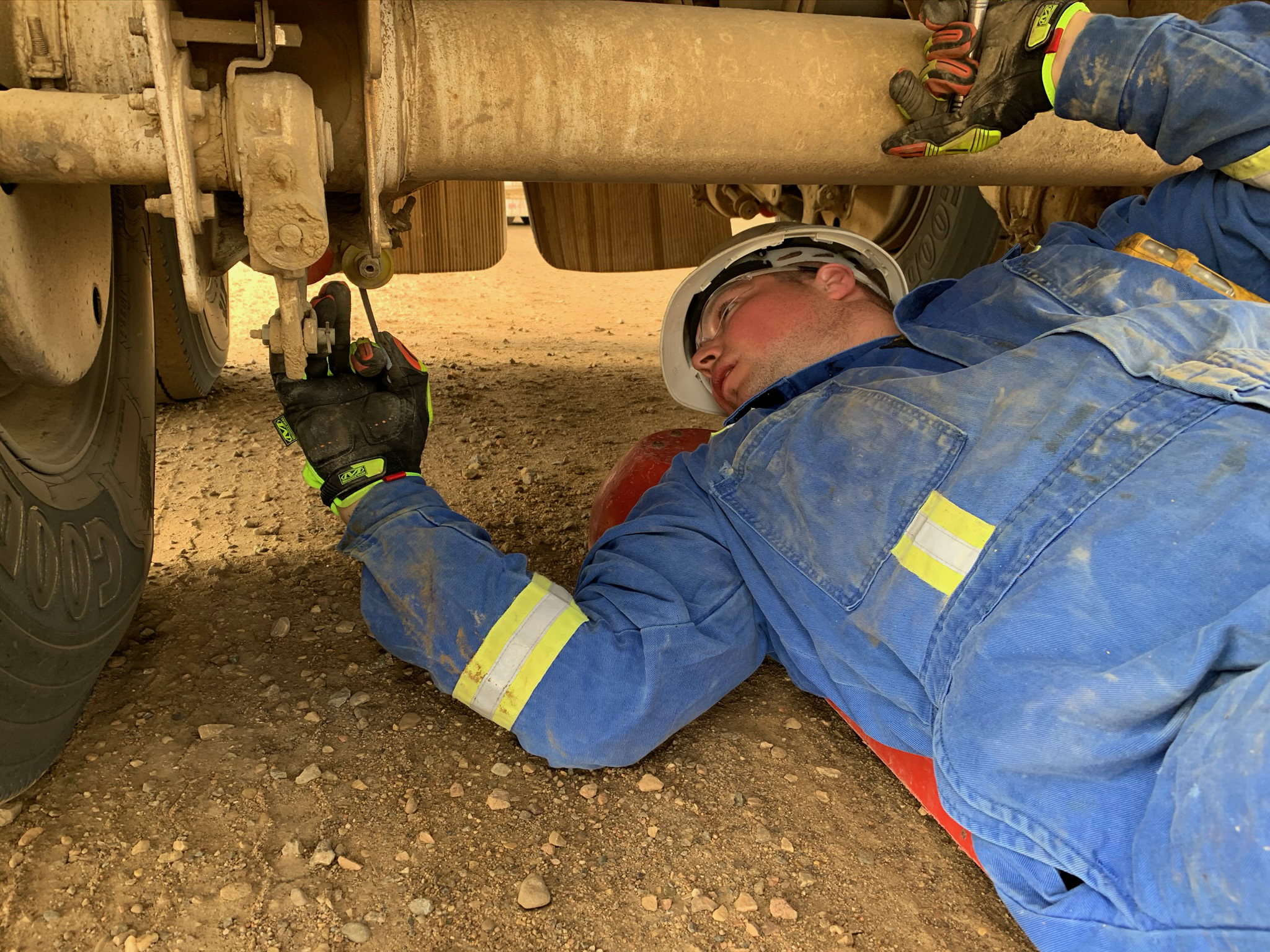 Mechanic in blue coveralls and hard hat working under a vehicle, performing maintenance on the suspension system.