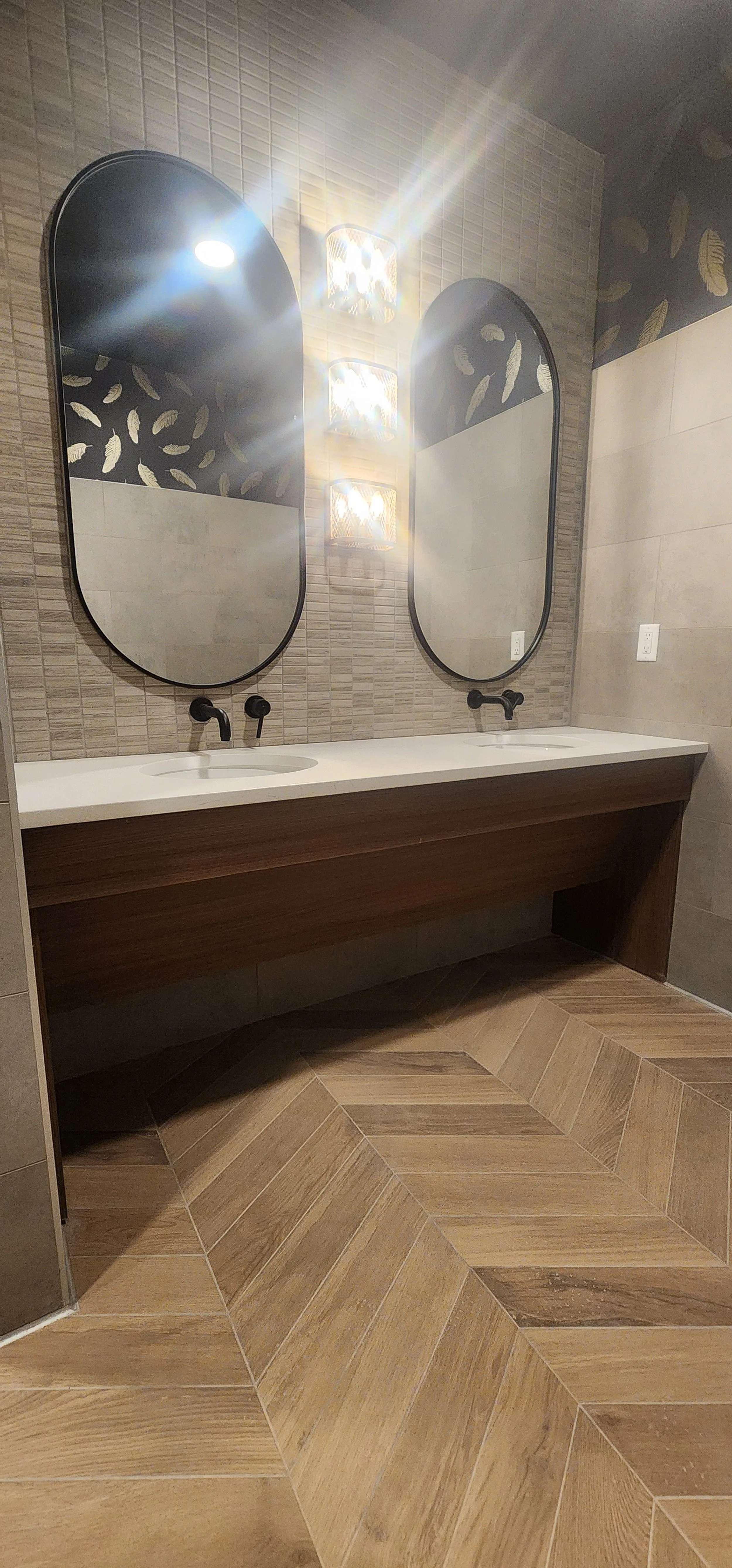 A modern bathroom with two oval mirrors above a white double sink vanity with black faucets. There is a wooden panel under the vanity, tile flooring in a herringbone pattern, and stylish wall lighting.