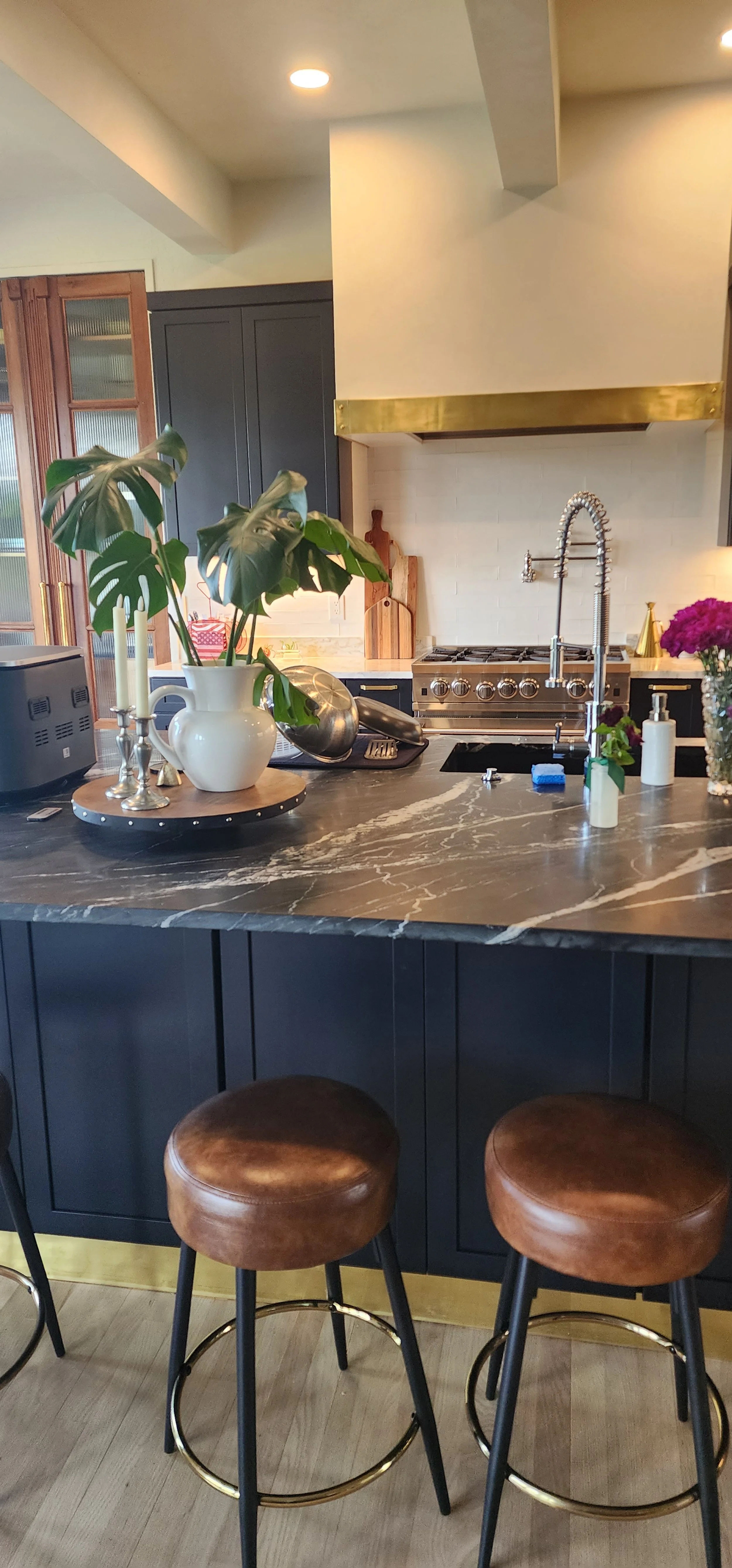 A modern kitchen with a stone countertop island, brown leather bar stools, and kitchenware on the counter, with black cabinetry and a gold range hood.