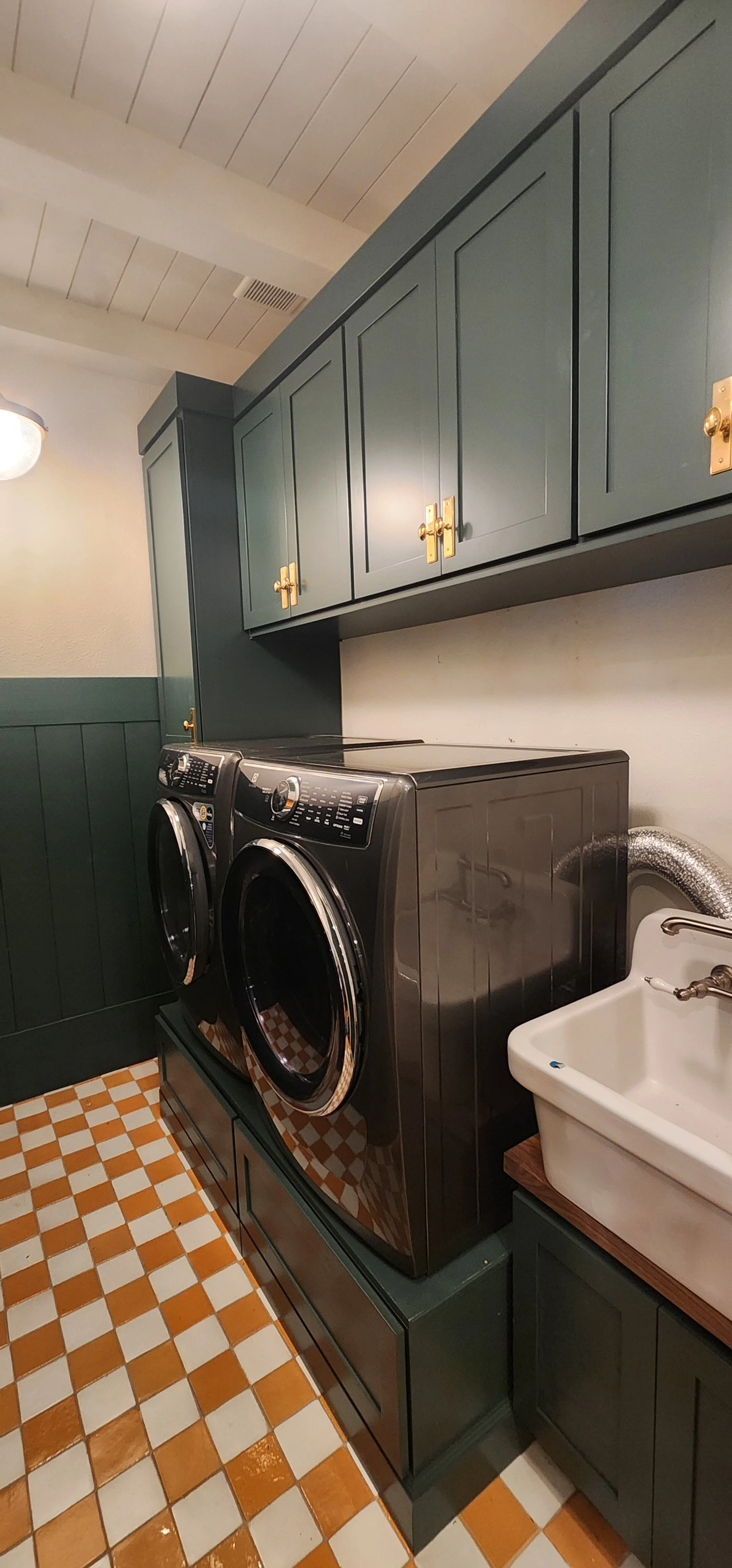 A laundry room with dark green cabinets, a white utility sink, and a black front-loading washing machine. The floor features orange and white checkered tiles.