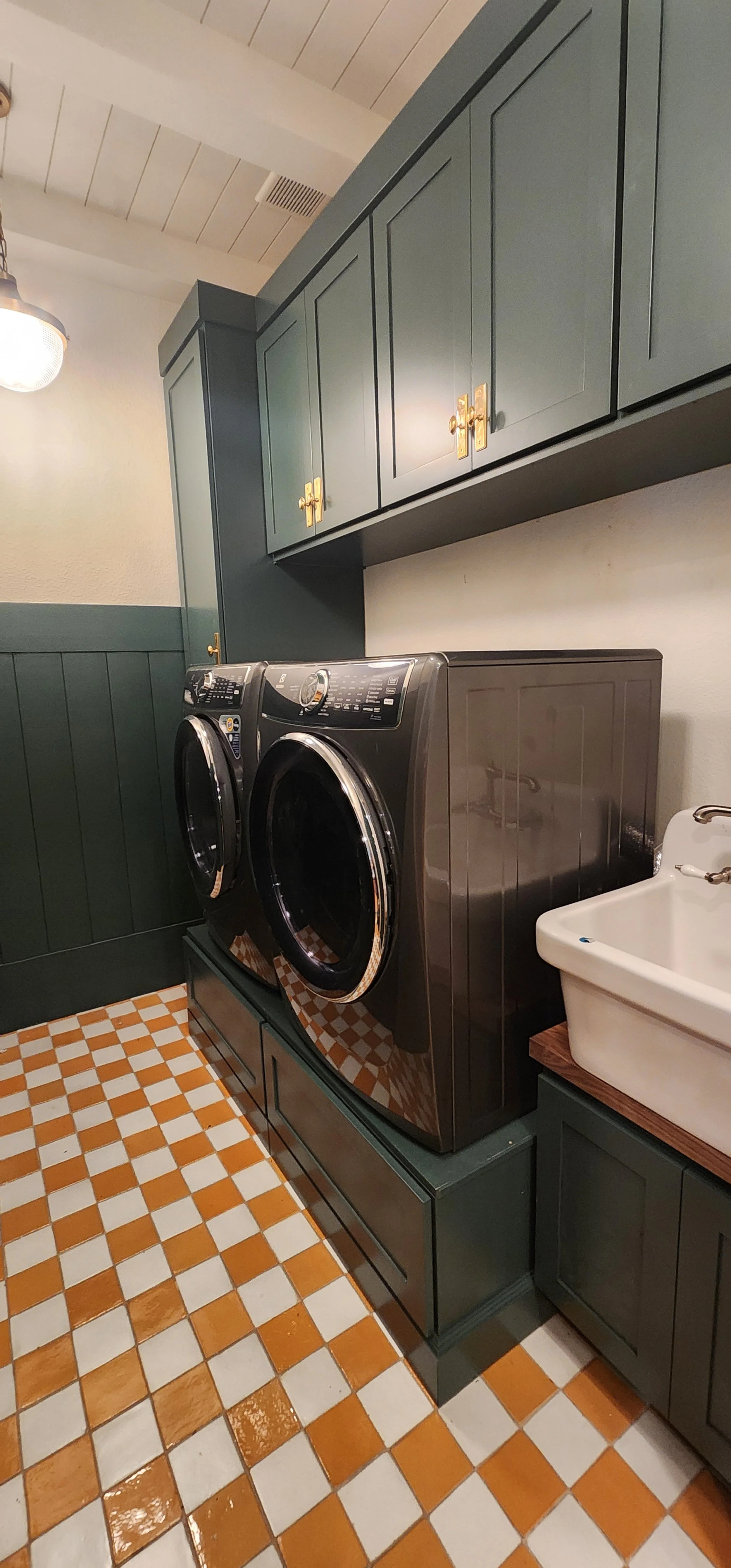 A laundry room with dark green cabinets with rolling washer and dryer pedestals, a front-loading washing machine and dryer, a white utility sink, and a tiled orange and white checkered floor.