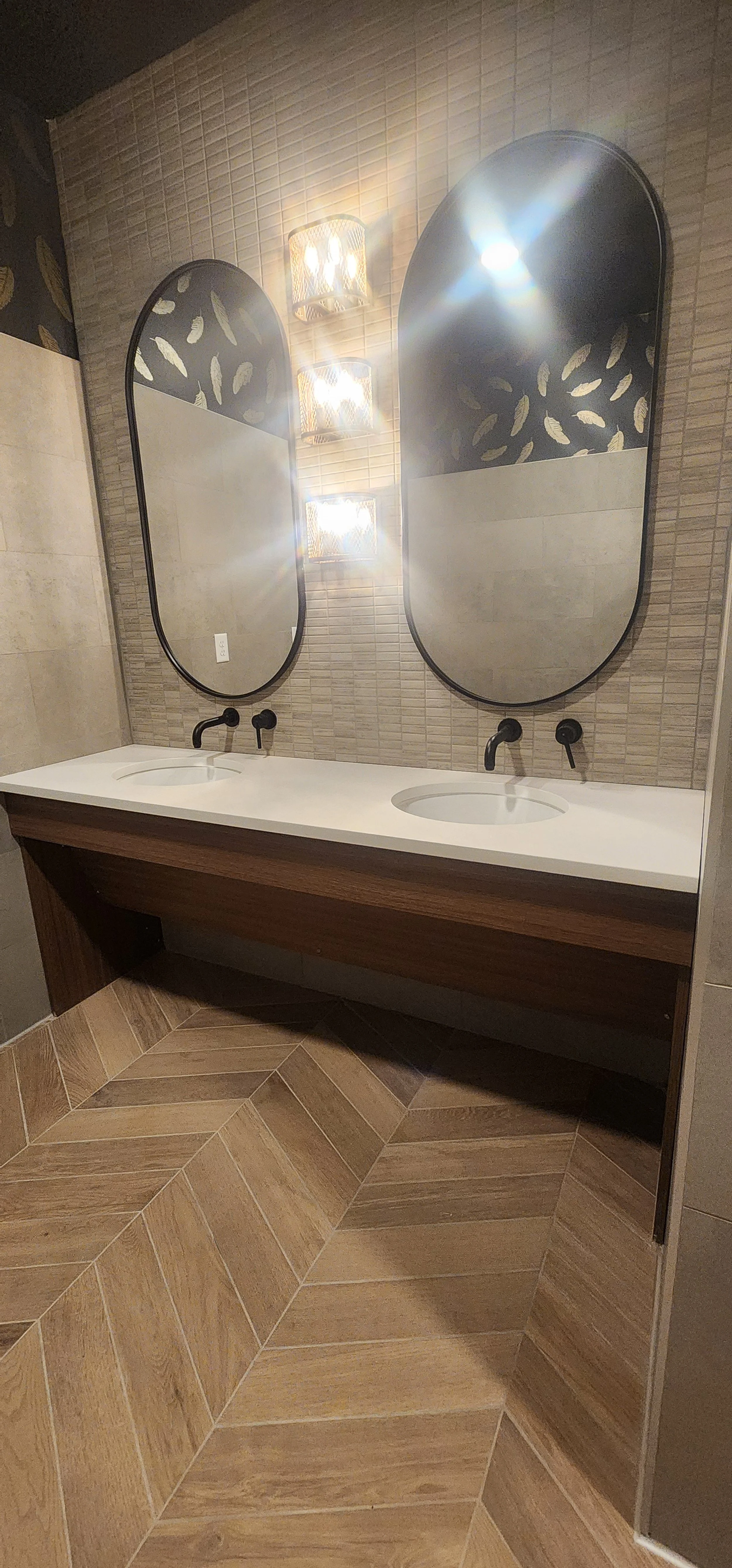 Modern bathroom with dual oval mirrors, wall-mounted black faucets, a double white countertop, colorful light fixtures, and wood-patterned tile flooring.