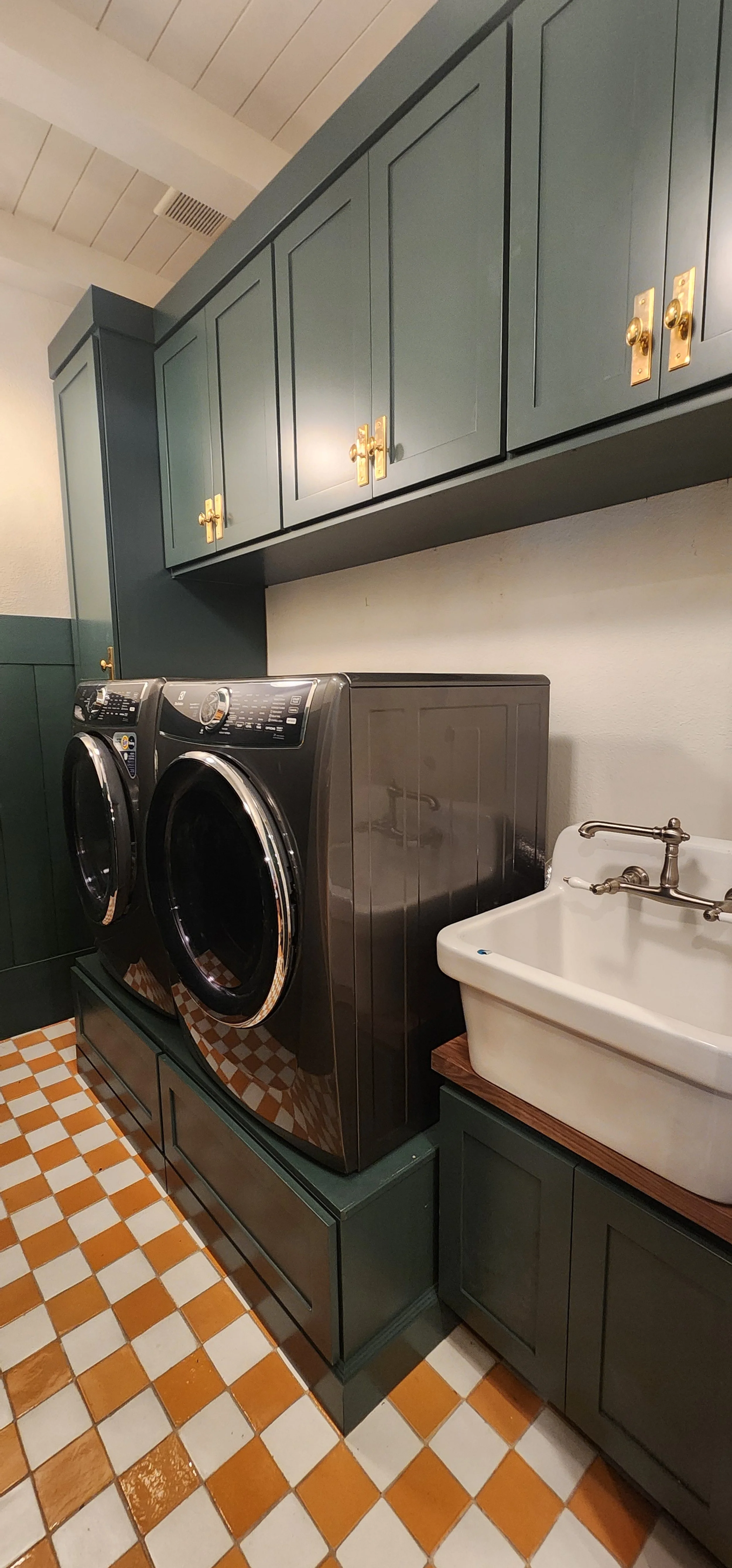 A laundry room with dark green cabinets, a front-loading washer and dryer, and a white utility sink with a brass faucet on a wooden countertop. The floor has orange and white checkered tiles.