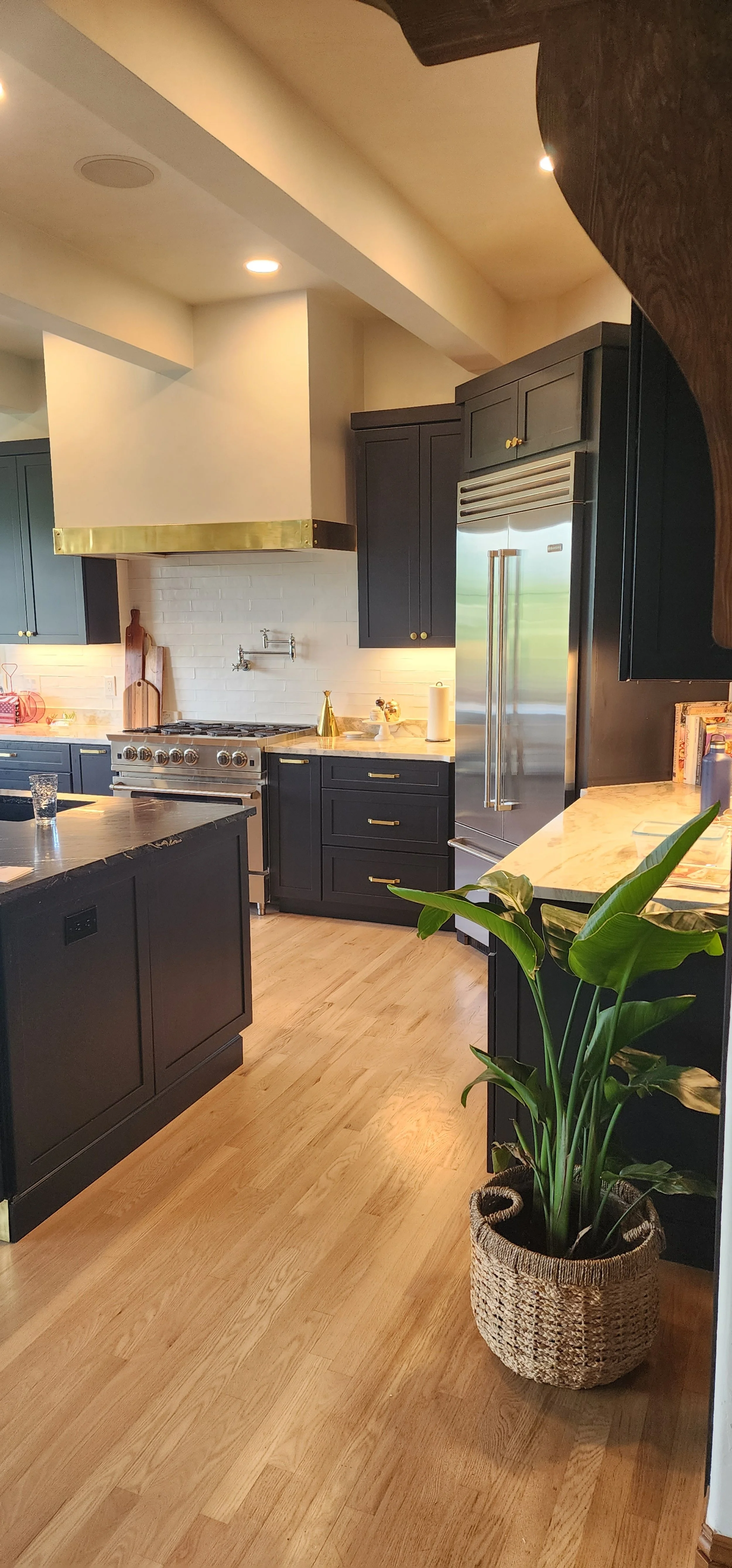 A modern kitchen with black cabinets, a Stainless-steel refrigerator, a white brick-style backsplash, and a wooden dining table. There is warm lighting from embedded ceiling lights.
