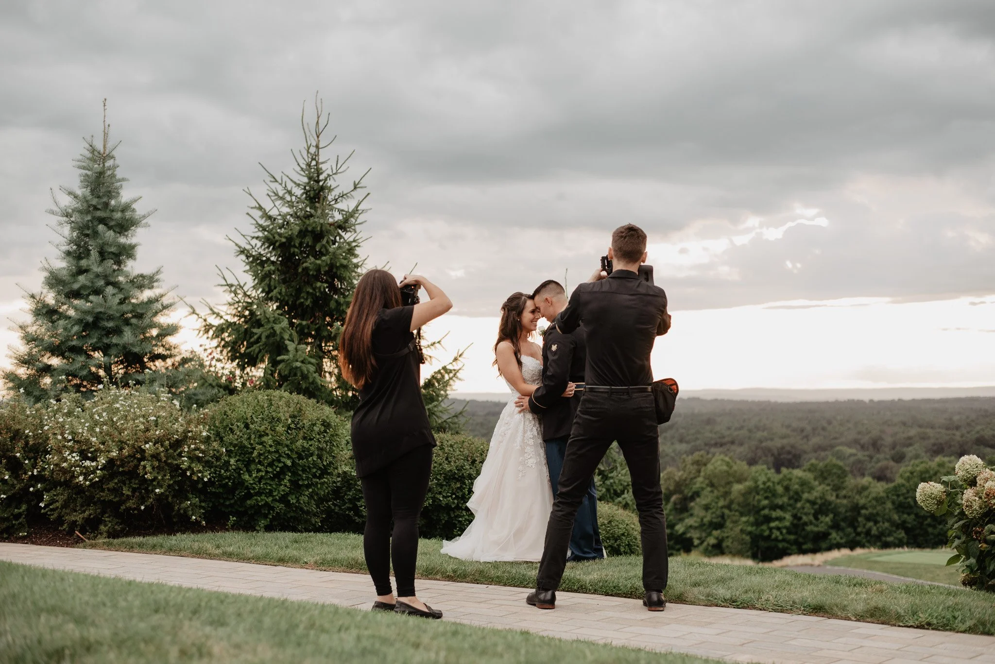 A couple getting married outdoors under cloudy skies, with two photographers capturing their moment among greenery and trees.