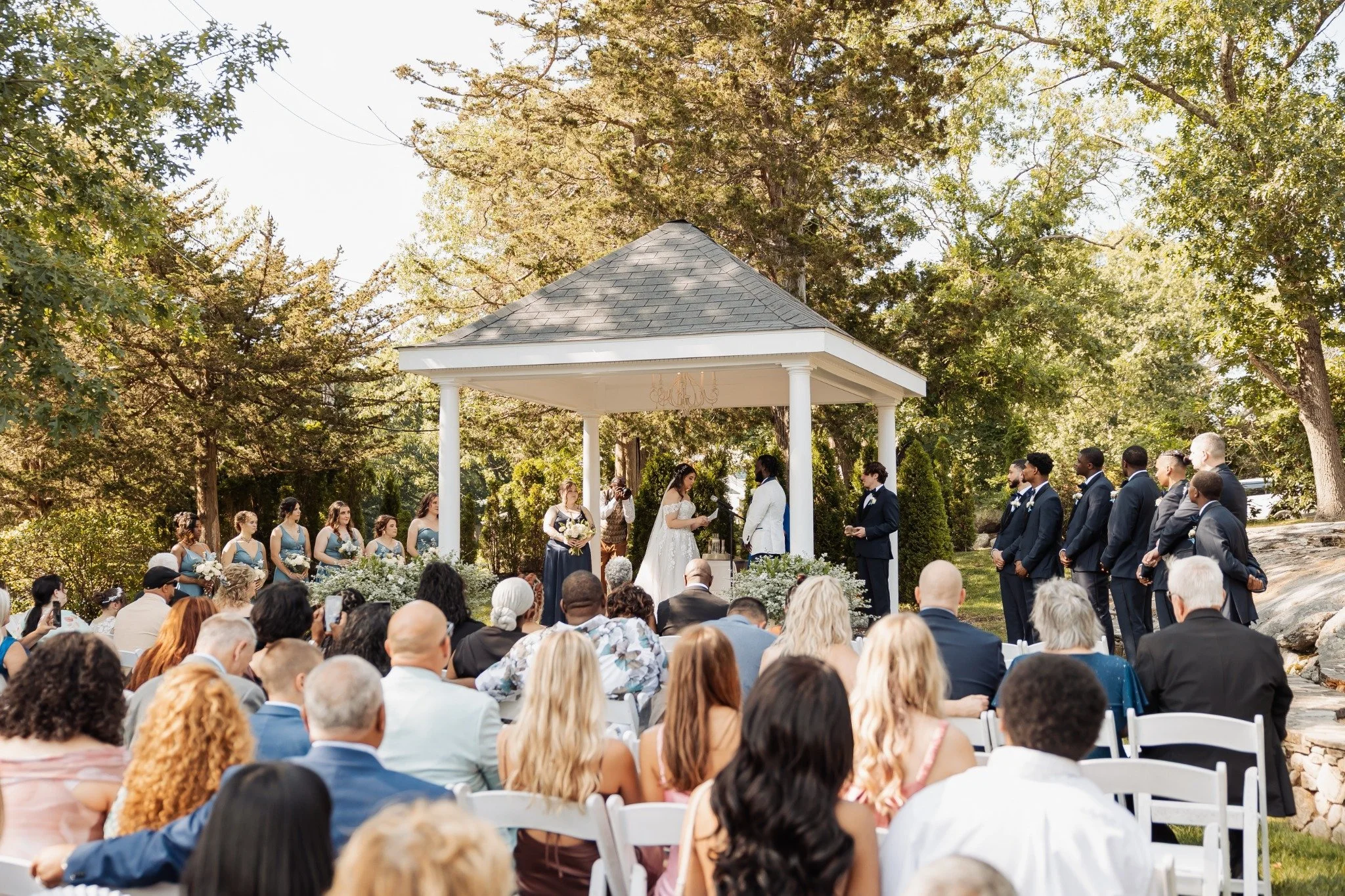 Outdoor wedding ceremony with bride and groom exchanging vows under a gazebo, surrounded by wedding party and seated guests, in a wooded area on a sunny day.