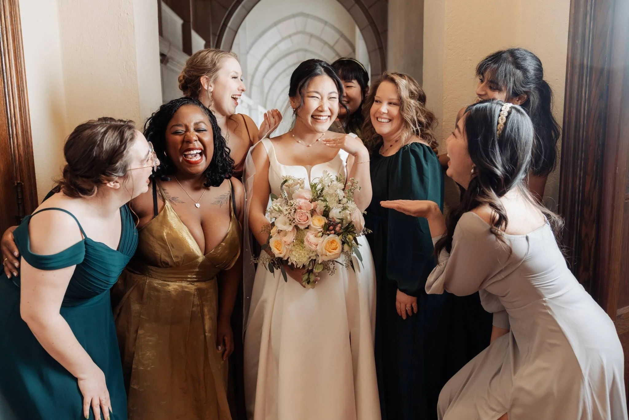 A bride in a white dress holding a bouquet of pink and white flowers surrounded by a group of women in colorful dresses, all smiling and laughing in an indoor setting.