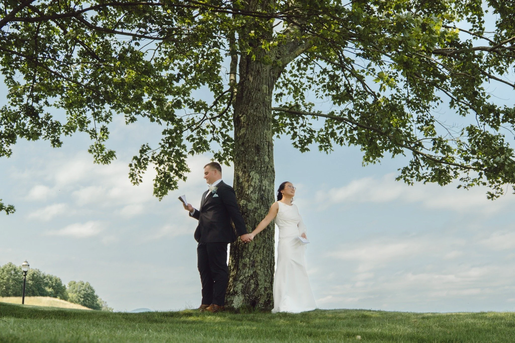A couple in wedding attire standing apart on either side of a large tree, holding hands, outdoors under a partly cloudy sky.
