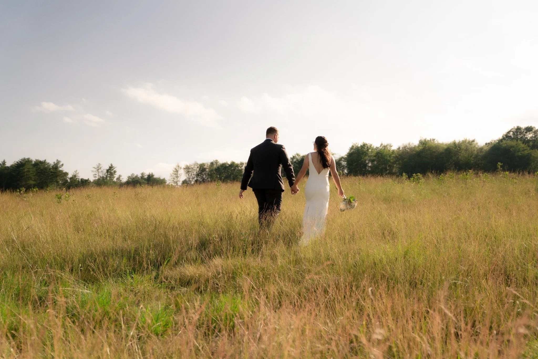 A bride and groom walking hand in hand through a grassy field on a sunny day, with the bride holding a bouquet.
