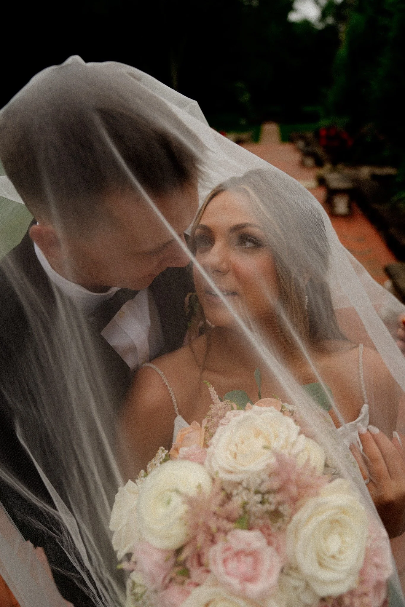 Bride and groom under a wedding veil, close-up of the couple with the bride holding a bouquet of white and pink flowers.