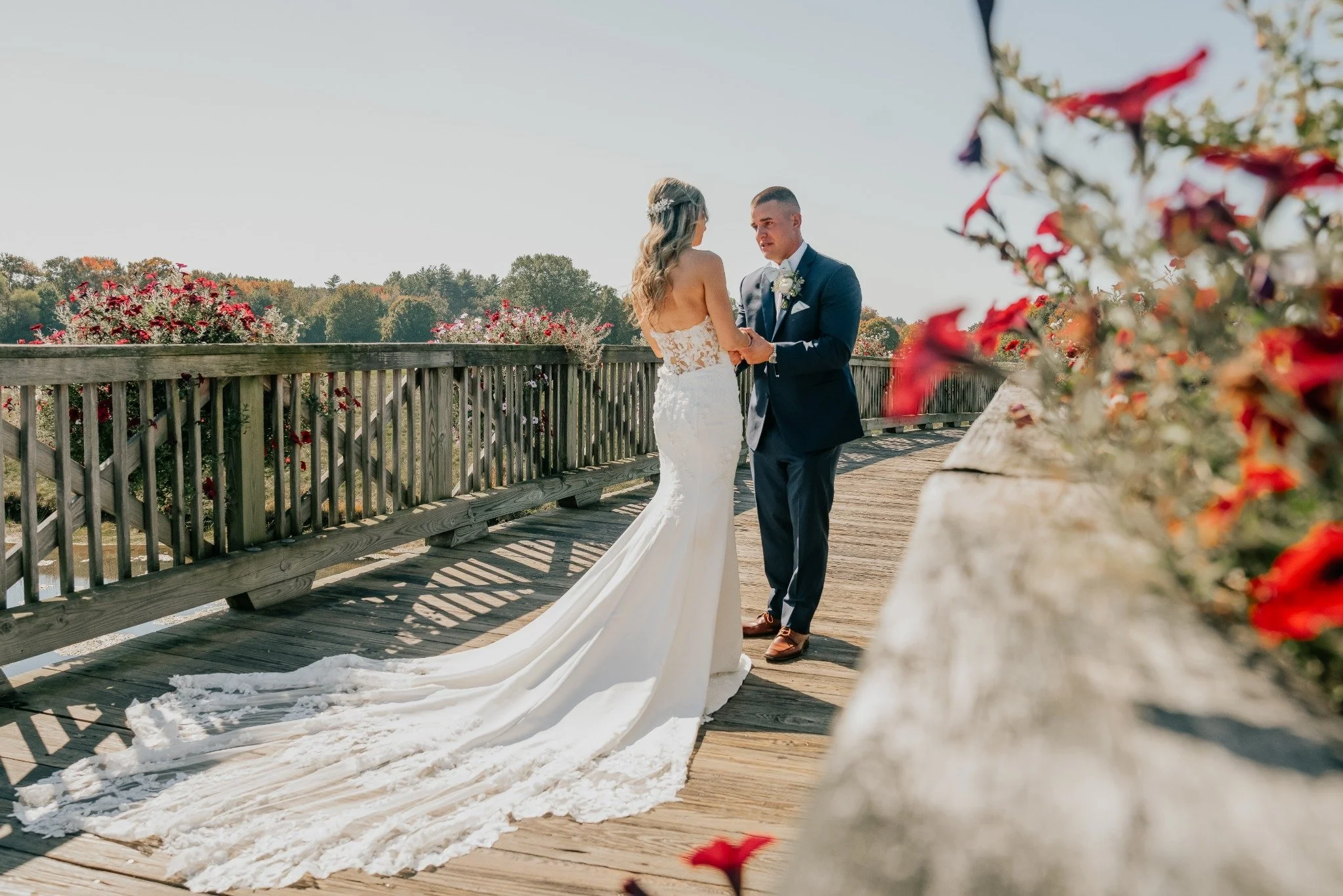 A bride and groom holding hands on a wooden bridge outdoors, with the bride in a white wedding dress with a long train, and the groom in a navy suit.