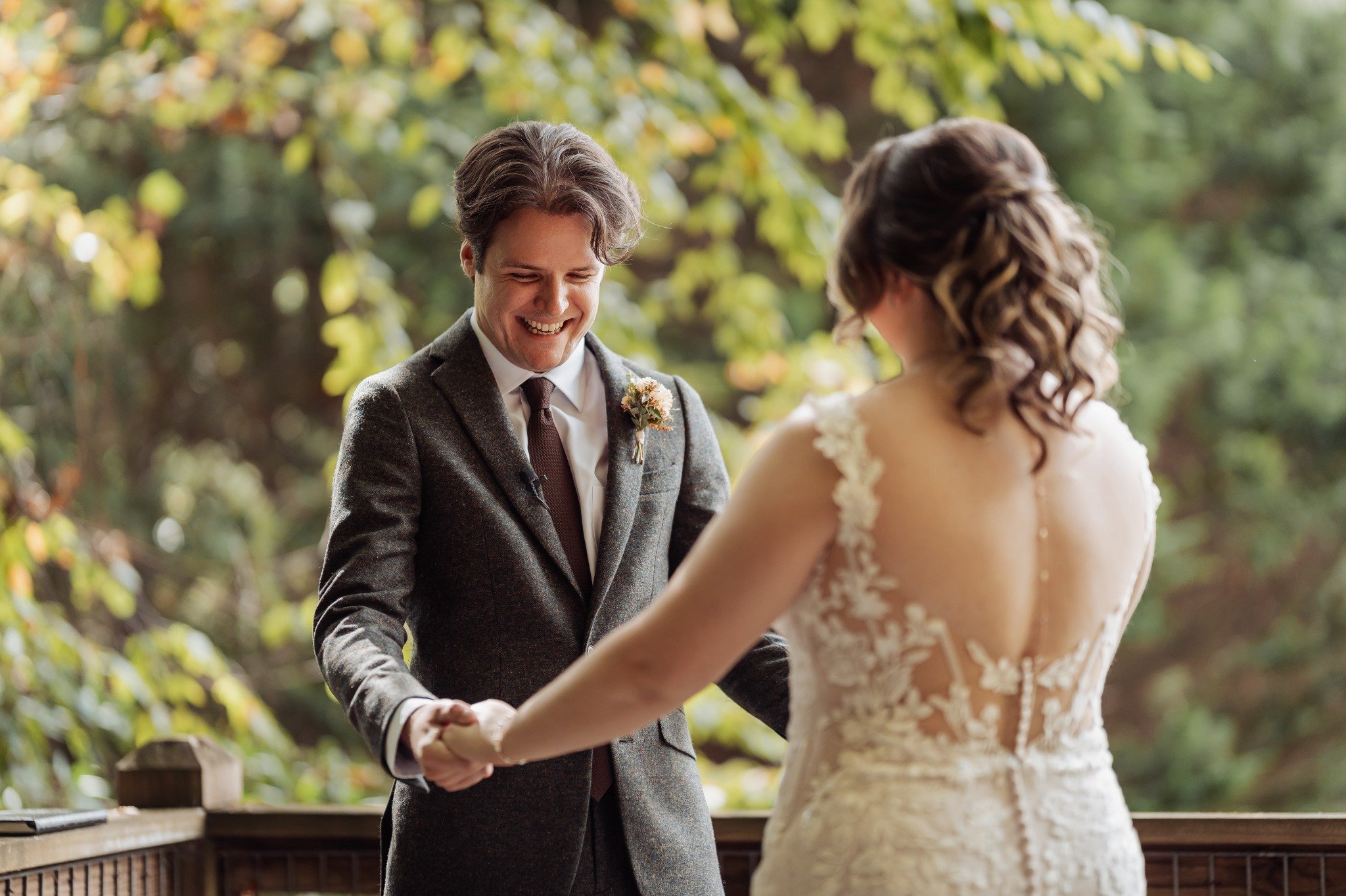 A couple exchanging wedding vows outdoors, the groom smiling and wearing a gray suit with a floral boutonniere, the bride in a white lace gown with a low back and curly hair, holding hands against a backdrop of green trees.