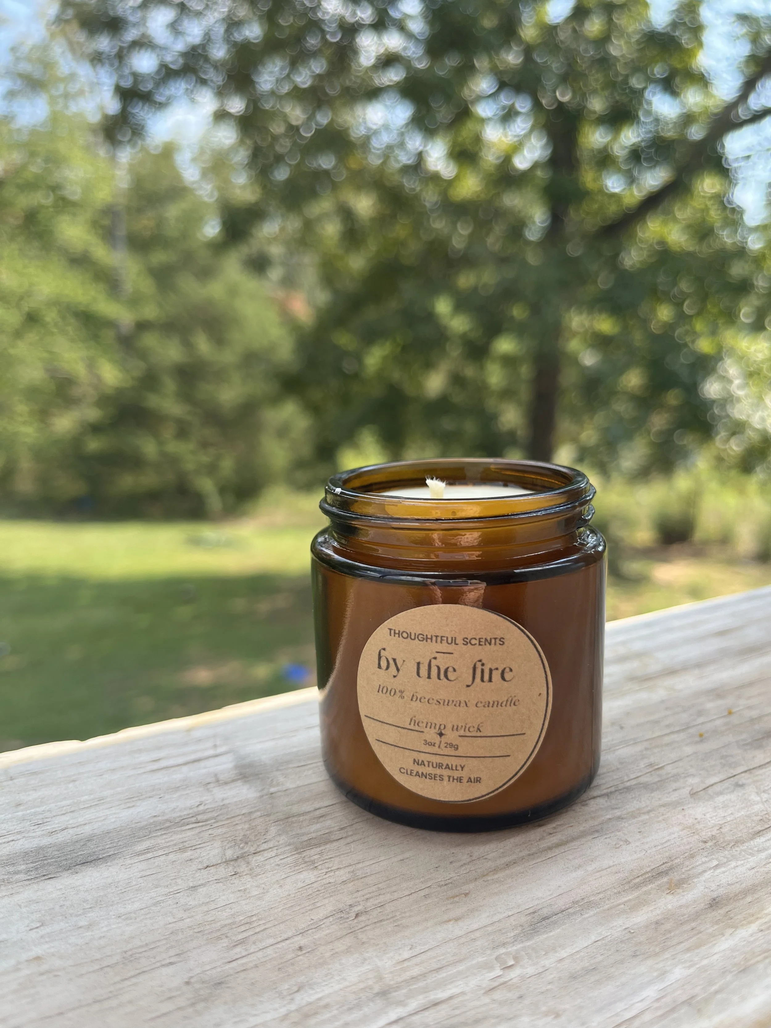 A brown glass jar candle with a white wax wick, placed on a wooden surface outdoors with green trees and a grassy area in the background.