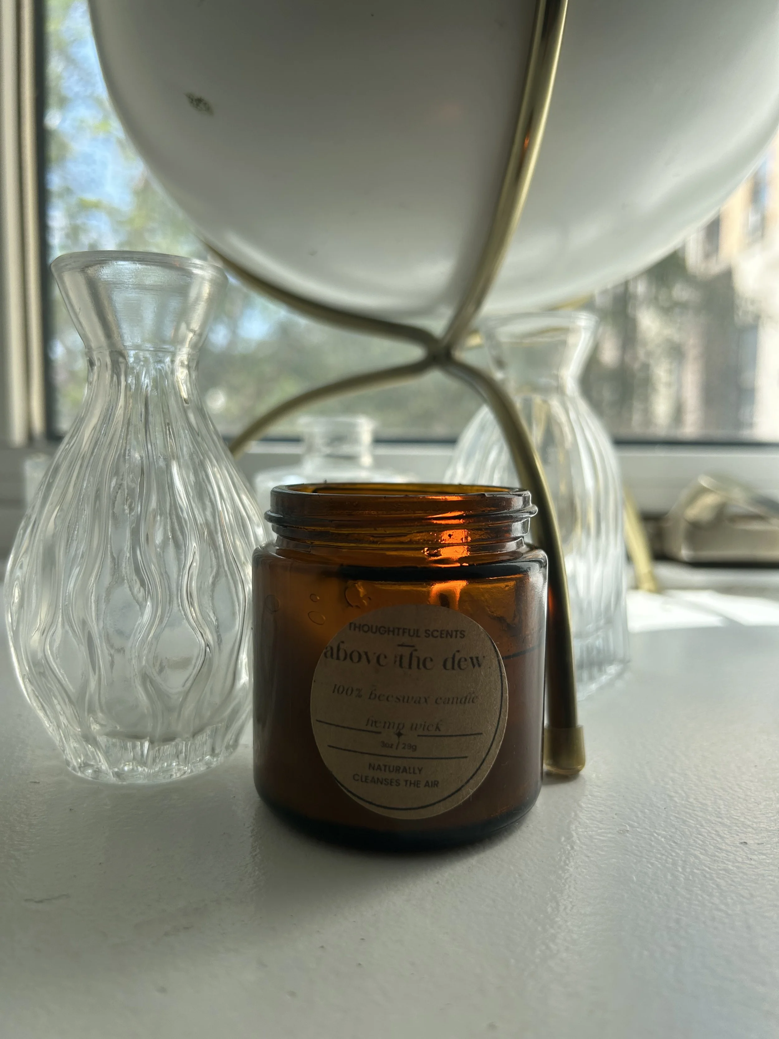 A brown candle jar labeled ‘above the dew,’ surrounded by three glass vases on a white windowsill, with a window overlooking trees in the background.