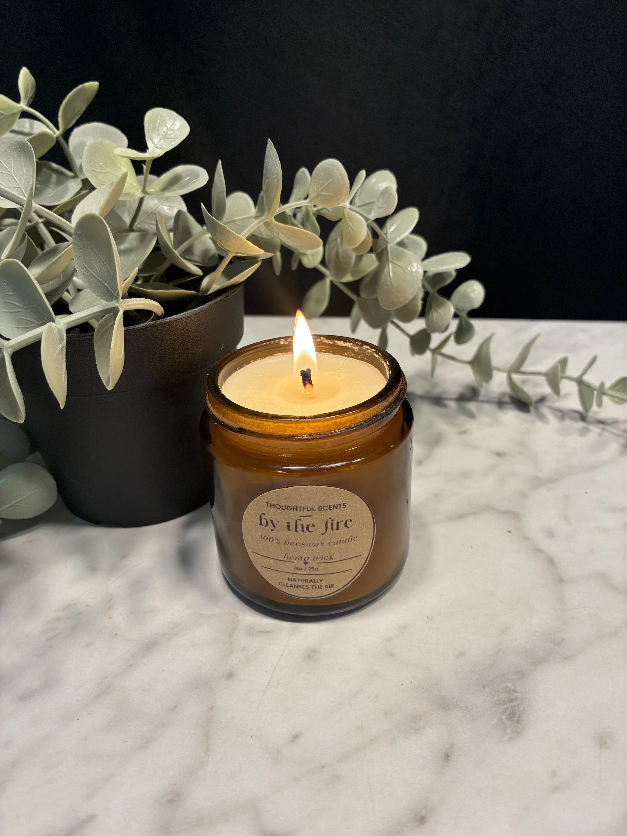 Lit candle in a brown glass jar with a label, placed on a white marble surface, next to a green leafy plant in a black pot, with a black background.
