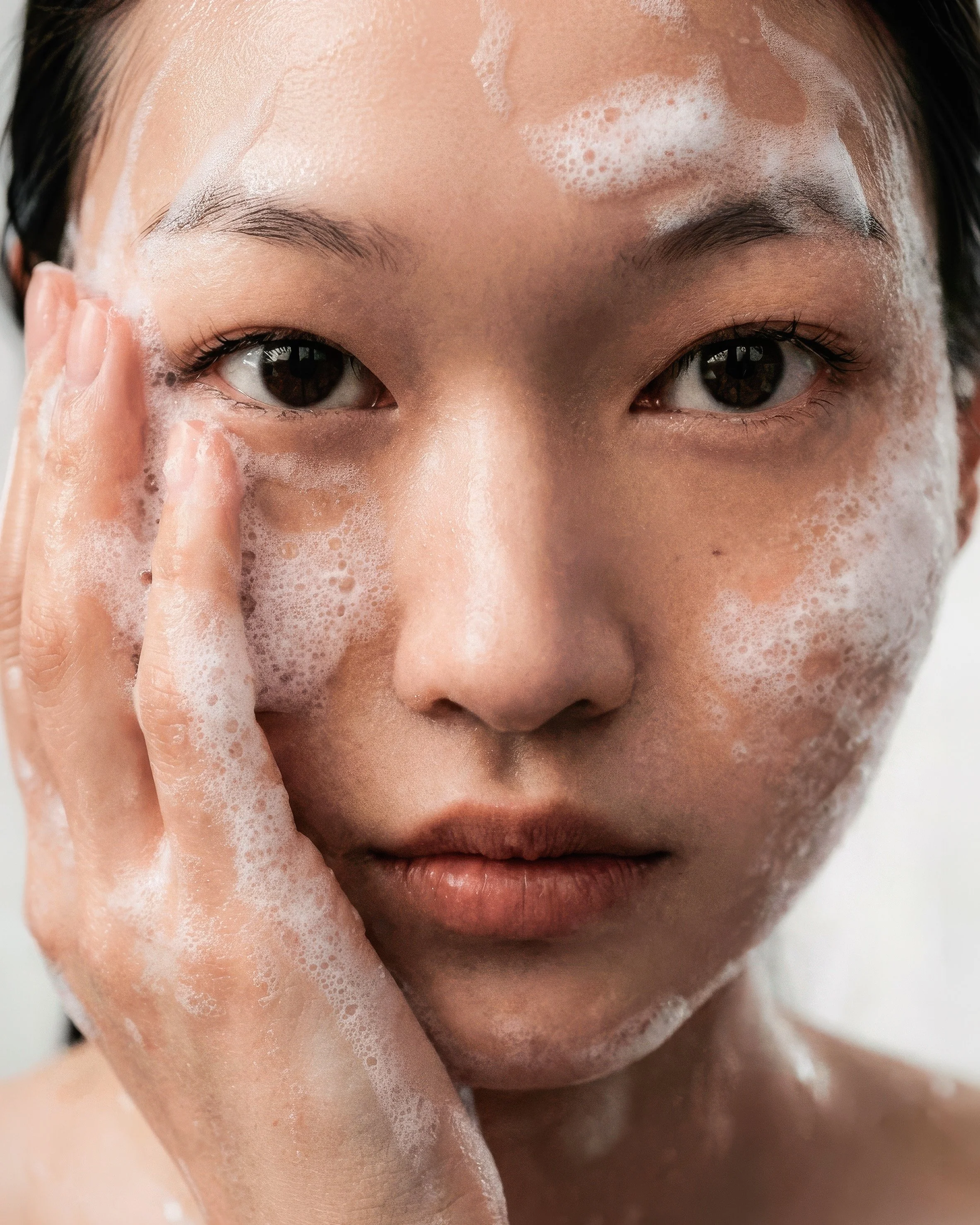 Close-up portrait of a woman gently washing her face with foamy cleanser, hand on cheek, dewy skin and soft natural lighting.