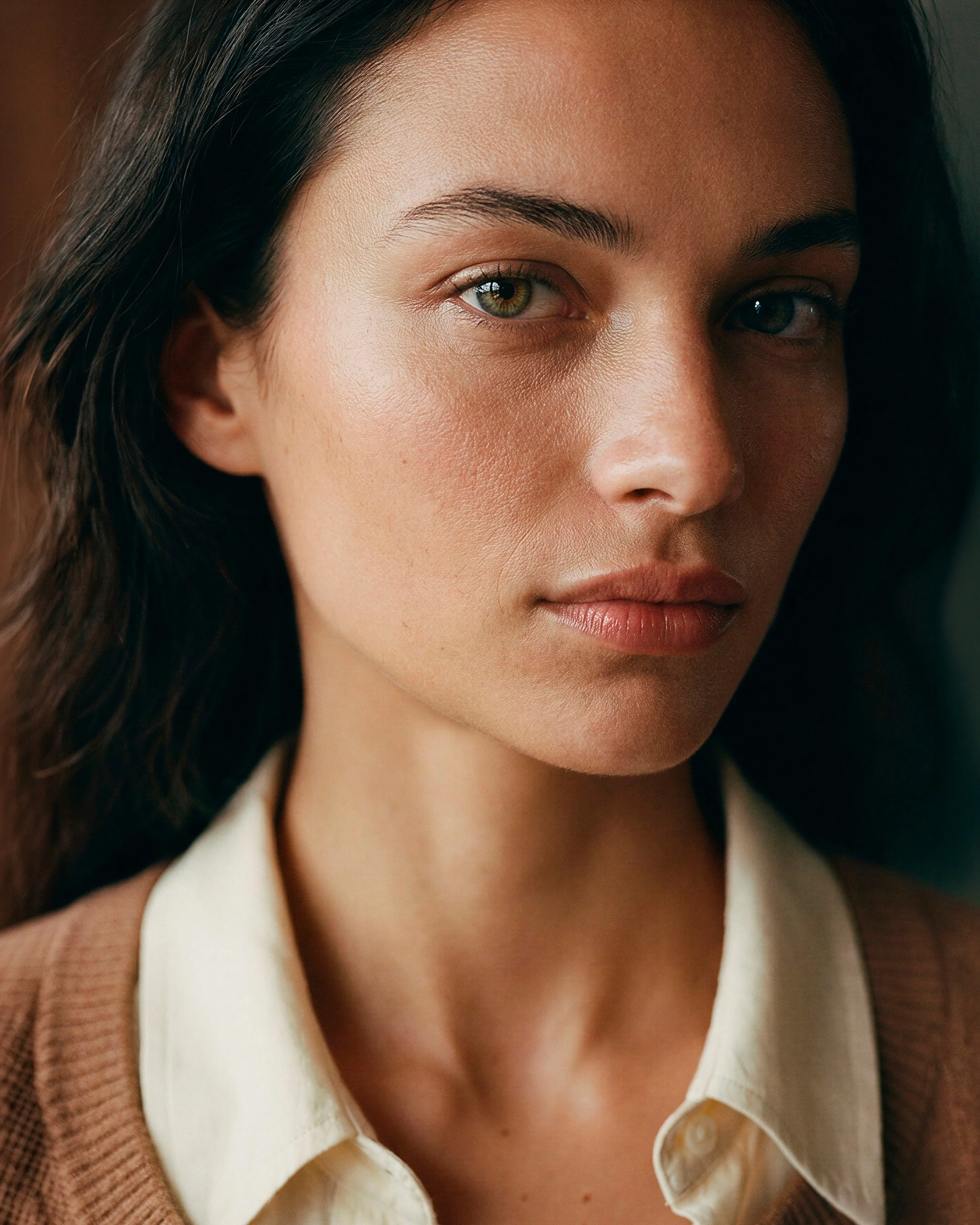 Close-up portrait of a woman with olive skin and dark hair, wearing a cream blouse and camel cardigan, softly lit with natural light against a warm, blurred interior background.