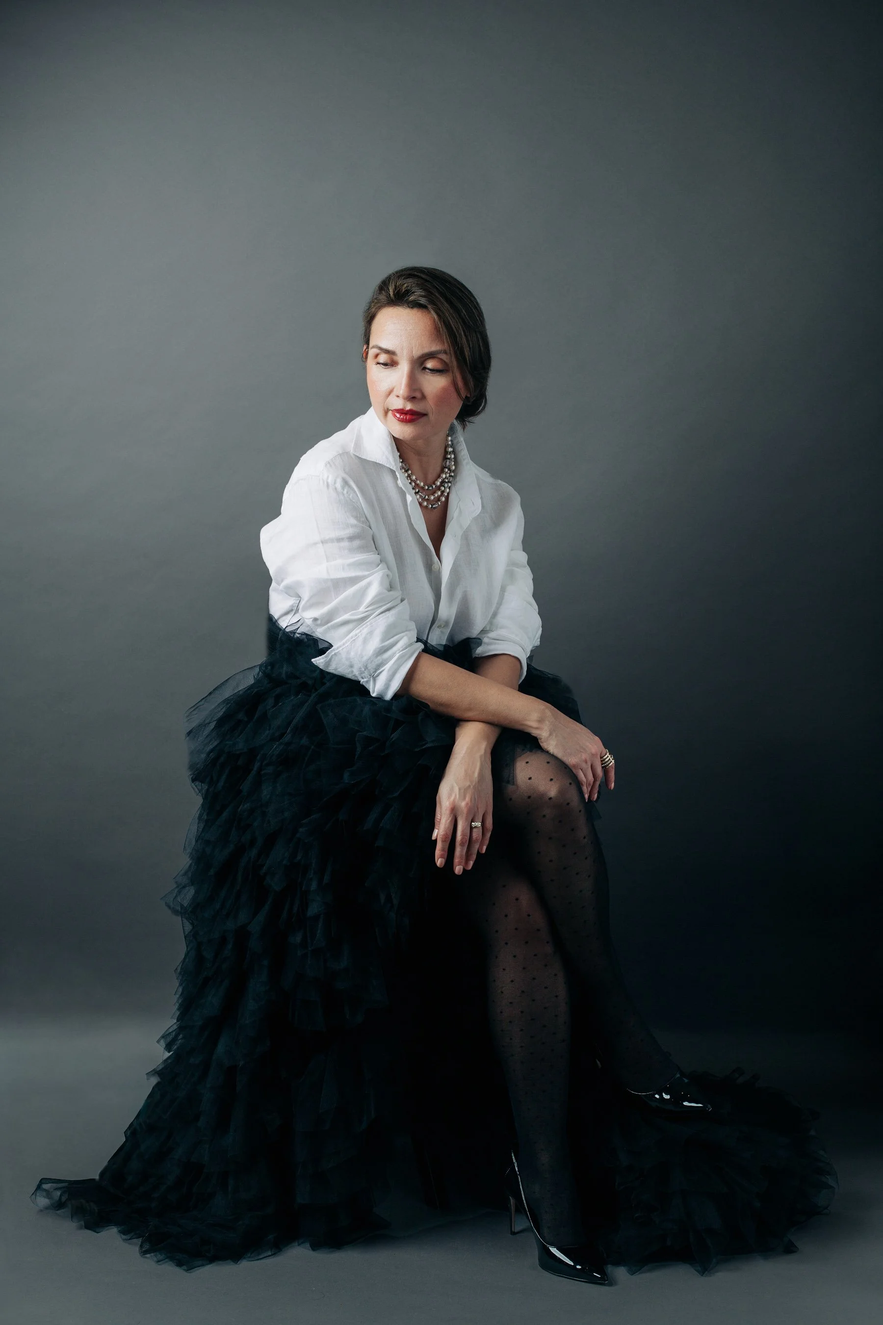 woman against gray backdrop, seated with her gaze down, remembering who she is, wearing a white blouse and black tulle skirt.