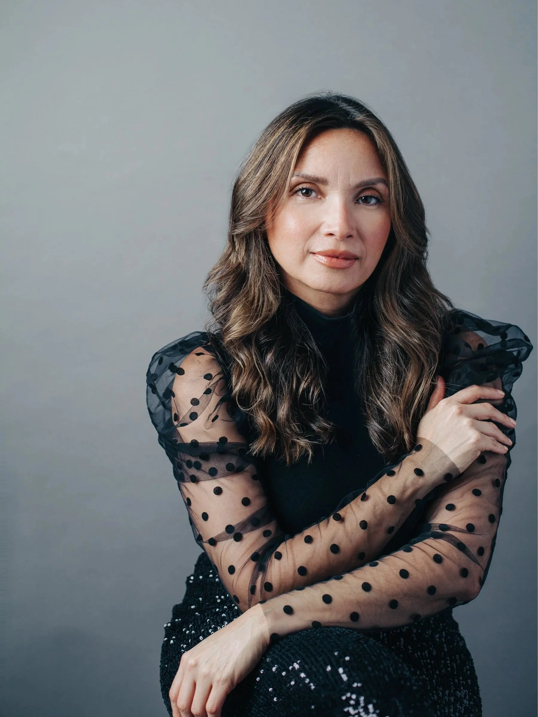 women seated against gray backdrop, one arm on her lap, one arm on her other arm, wearing black shear polka dot blouse and sequin skirt, long wavy hair, soft smile, direct eye contact.