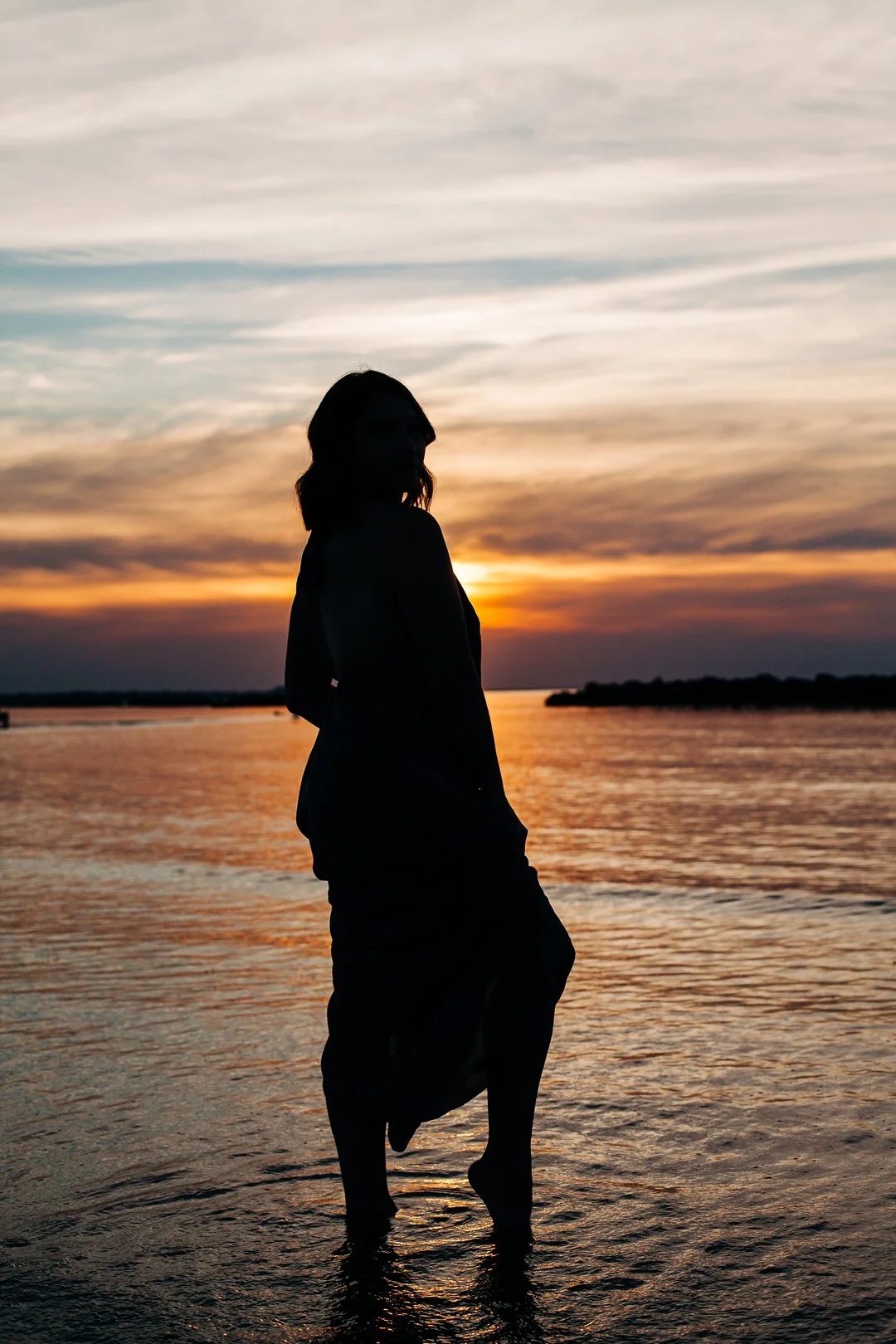 Silhouette of a woman standing beside calm water at sunset.