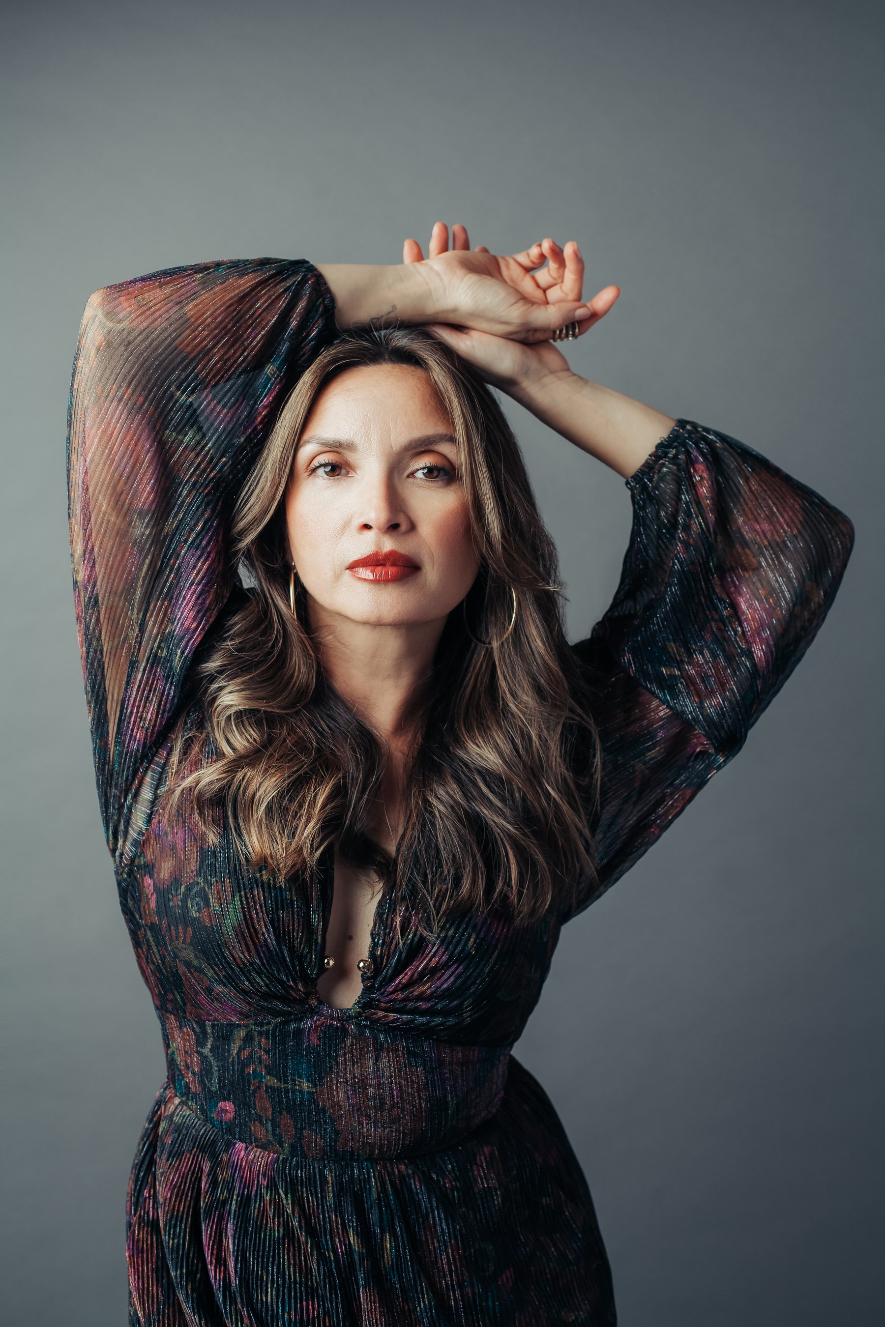 Woman against gray backdrop, she's wearing a long sleeve floral dress, her hands are above her head, her gaze direct, she embodies presence.