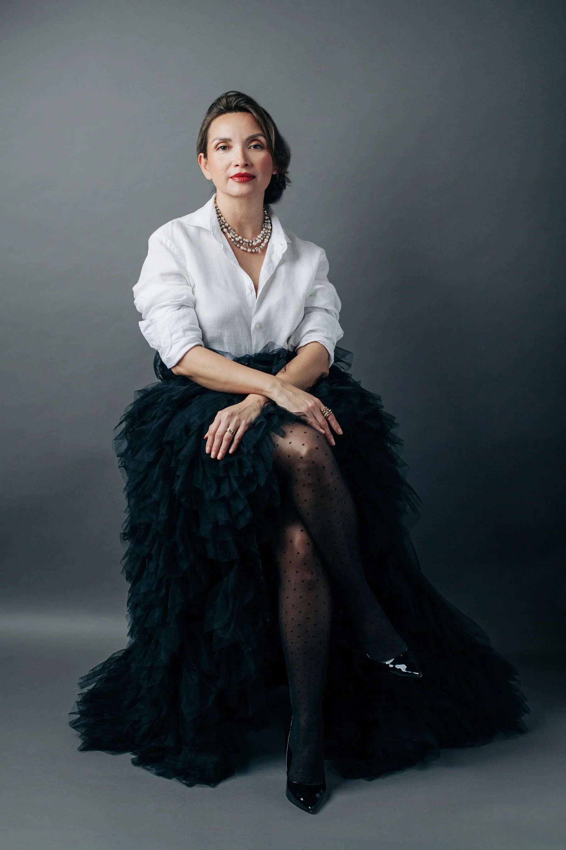 Woman seated against a gray backdrop, wearing a white blouse and a black tulle skirt, sitting upright with a calm posture and a gaze, embodying grounded confidence and quiet authority.