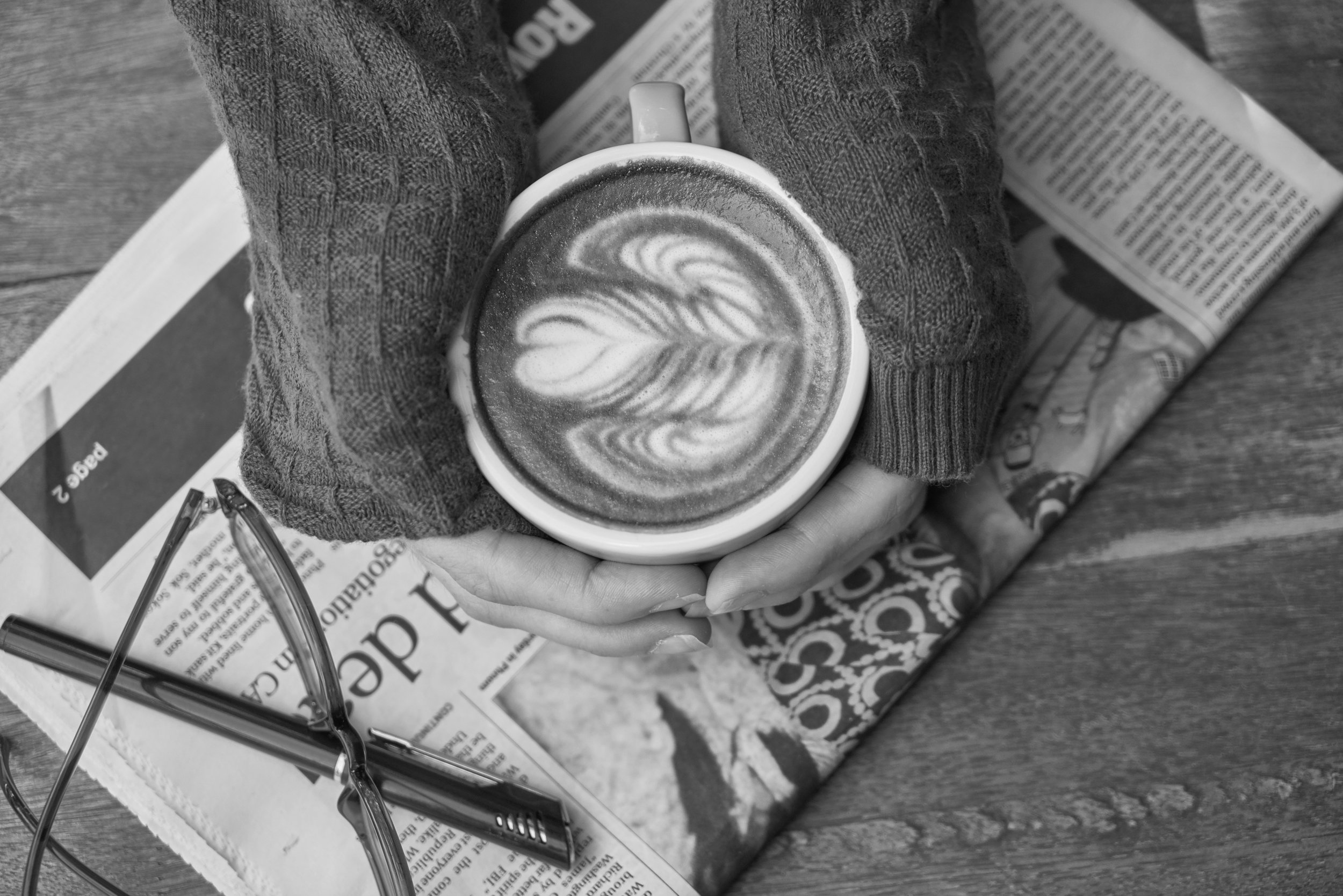 Two hands cupping a latte, resting atop a newspaper, seen from above.